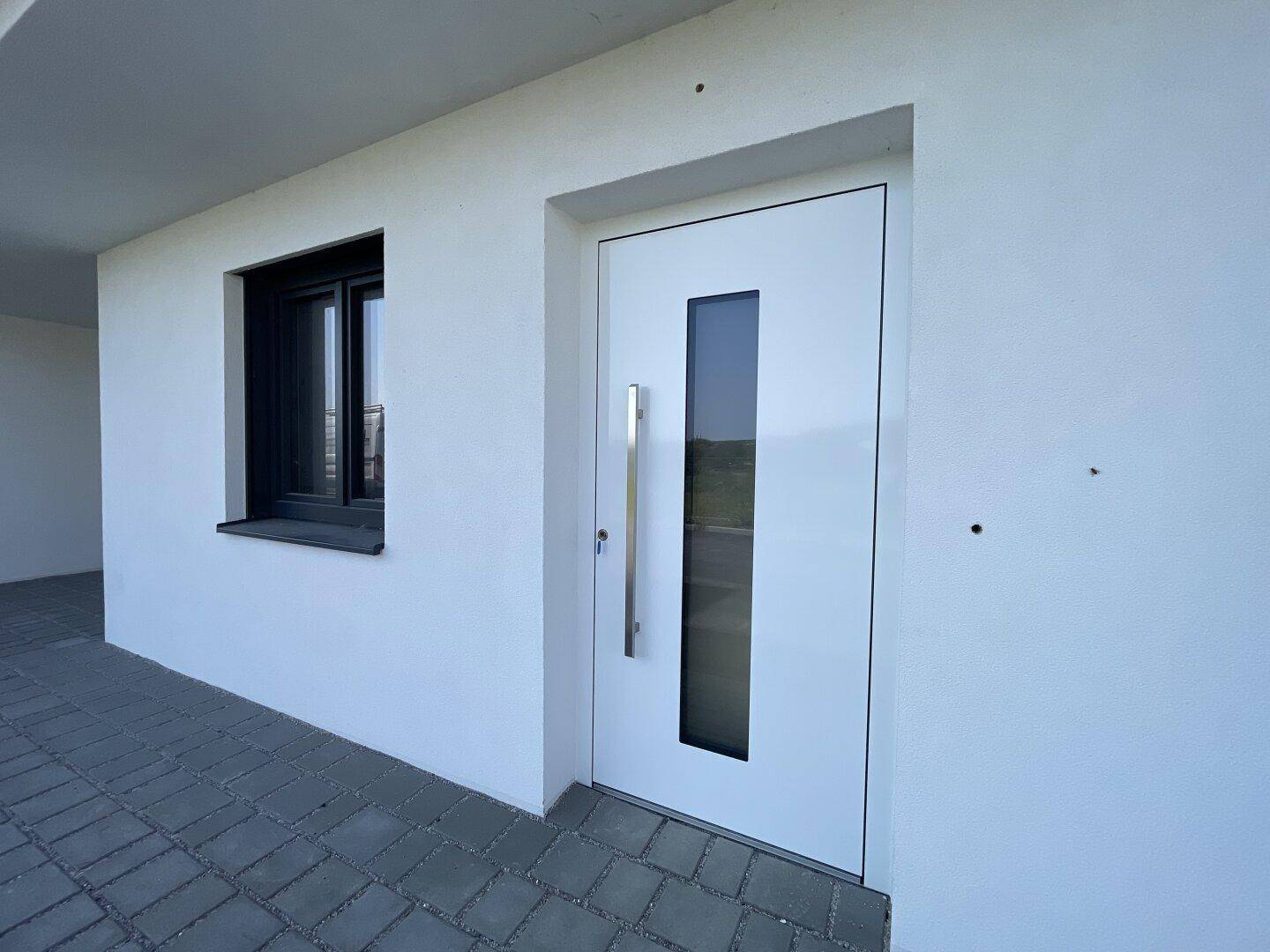 Weiße Eingangstür mit vertikalem Glaseinsatz und modernem Griff, umgeben von heller Fassade.