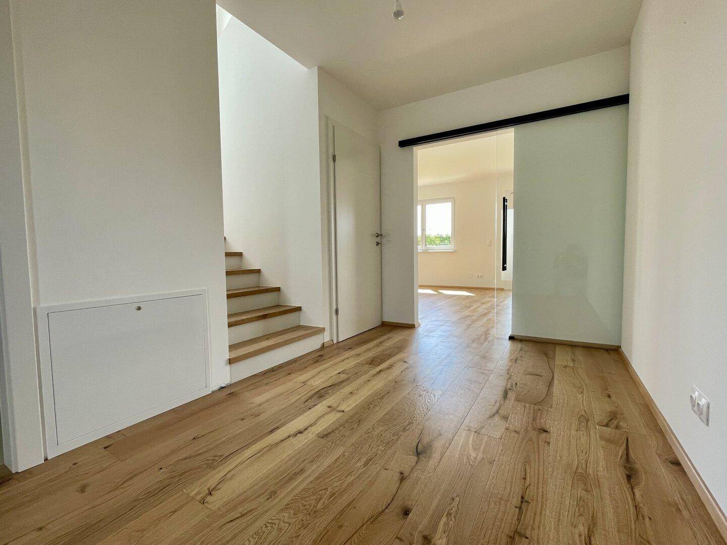 Heller Flur mit Echtholzdielenboden, moderner Treppe und Glasschiebetür zu den Wohnbereichen.