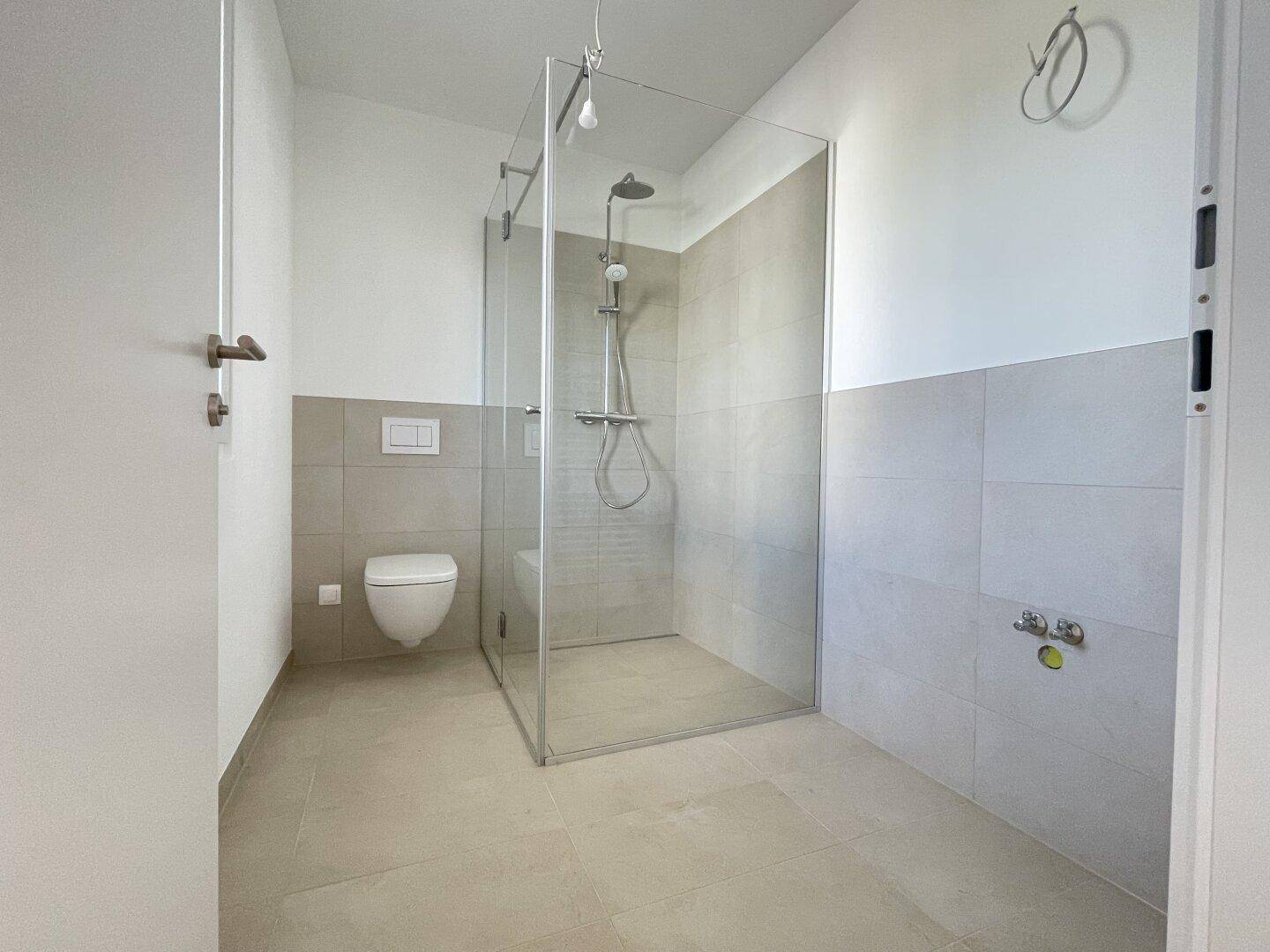 Modernes Badezimmer mit hellgrauen Fliesen, wandhängendem WC und geräumiger begehbarer Dusche.