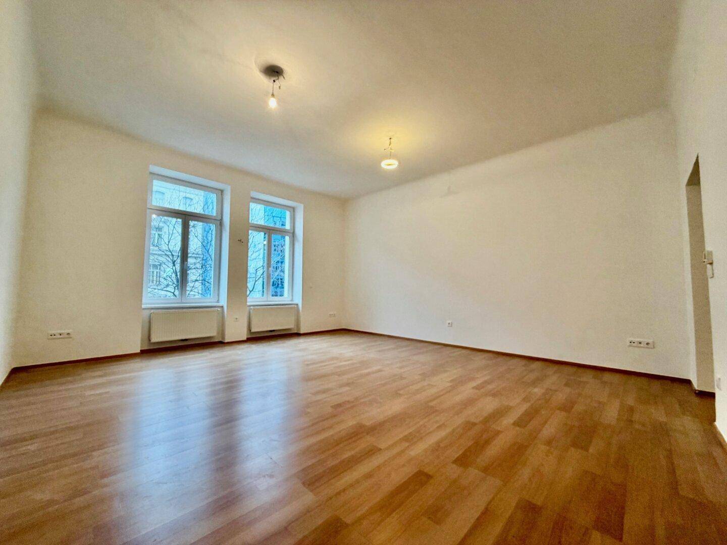 Geräumiges, leeres Wohnzimmer mit hellem Holzboden und zwei großen Fenstern, die viel Tageslicht hereinlassen.