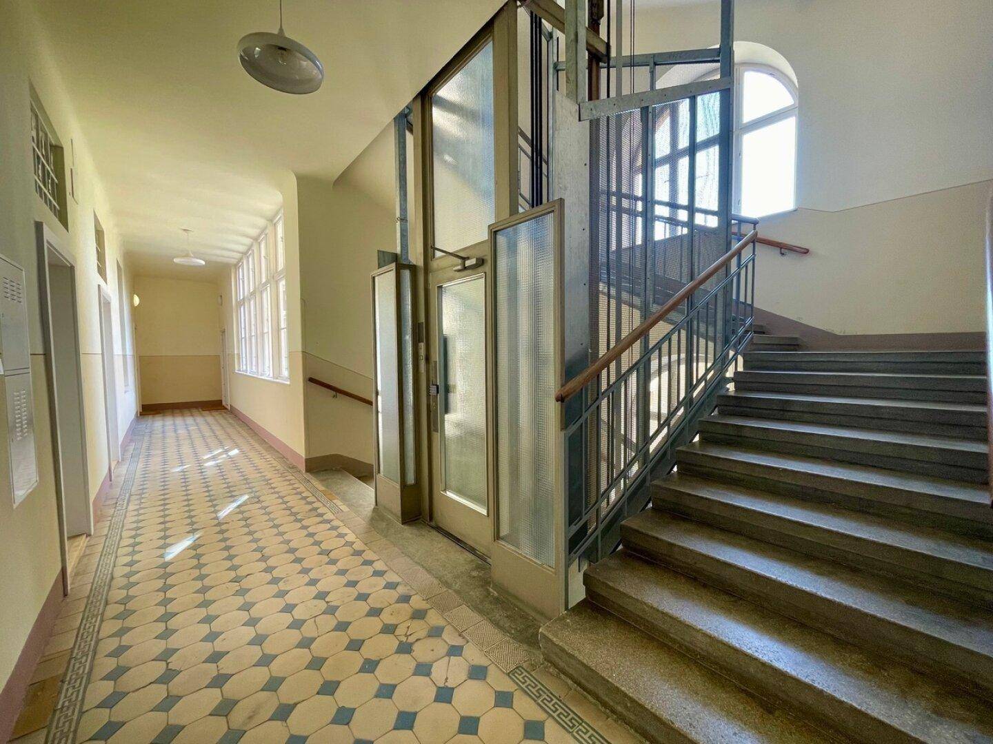 Historisches Treppenhaus mit Aufzug, kunstvollen Fliesen und großen Fenstern, die den Gang erhellen.