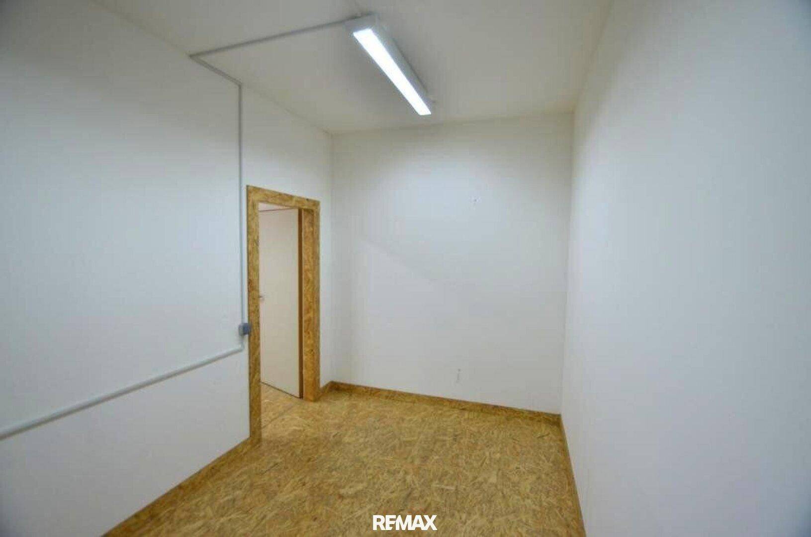 Kleiner Innenraum mit hellen Wänden, OSB-Boden und einer Tür, die zu einem weiteren Raum führt.