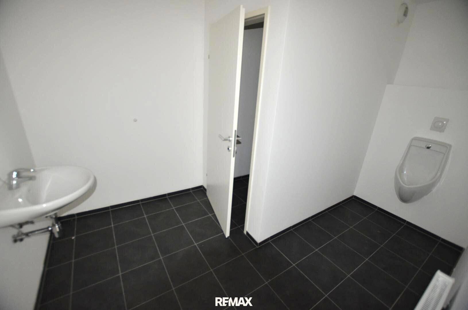 Kleiner Toilettenraum mit Waschbecken, Urinal und dunklen Bodenfliesen, funktional und sauber.