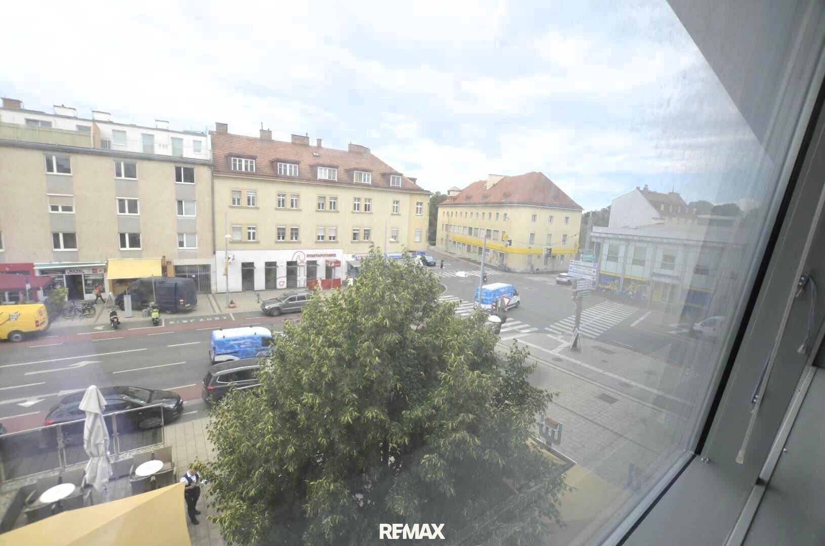 Blick aus dem Fenster auf eine belebte Stadtstraße mit Gebäuden, Fahrzeugen und Bäumen.