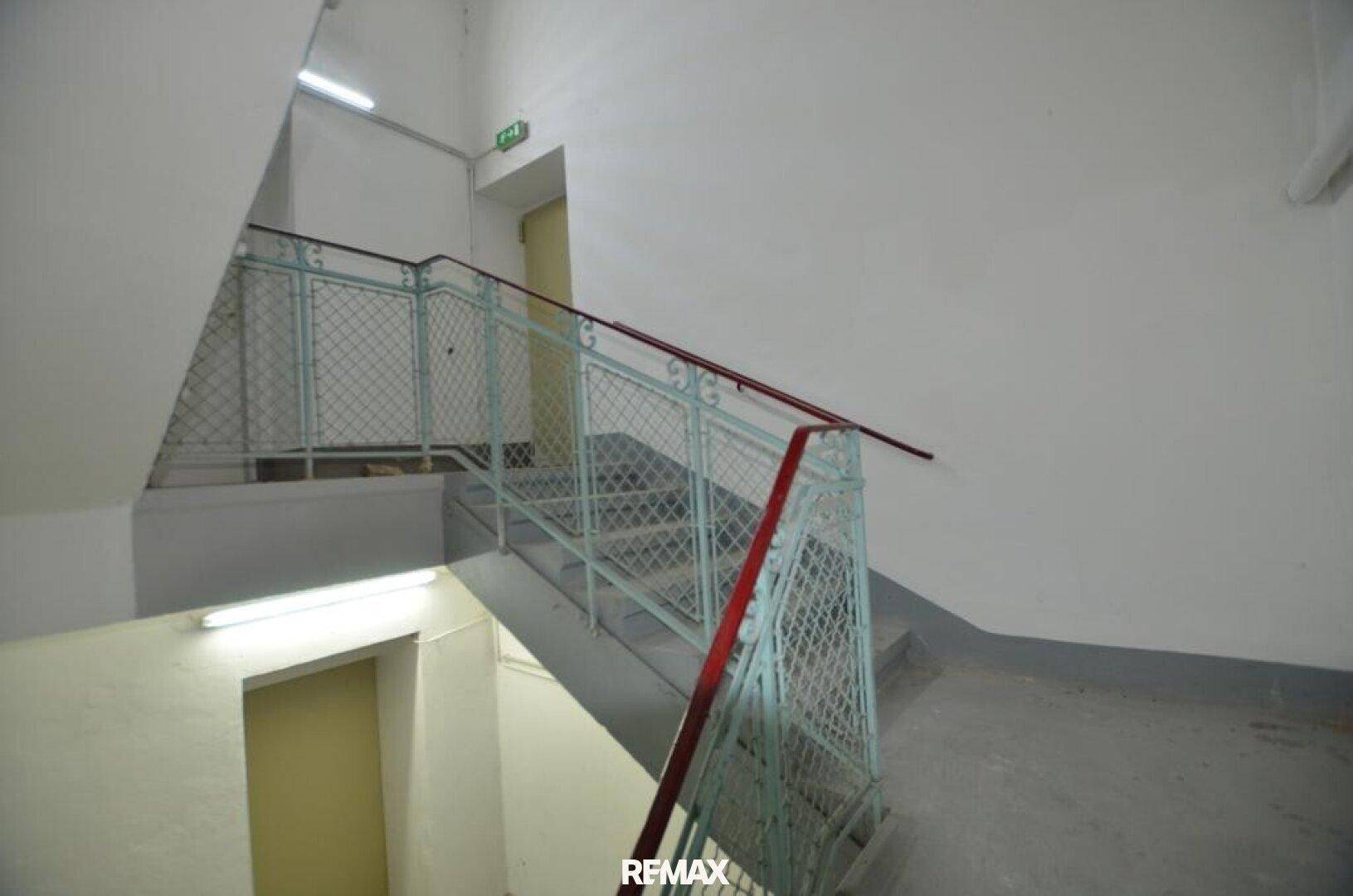 Treppenhaus mit Metallgeländer und Betonstufen, beleuchtet von Leuchtstoffröhren, mit Ausgangsschild.