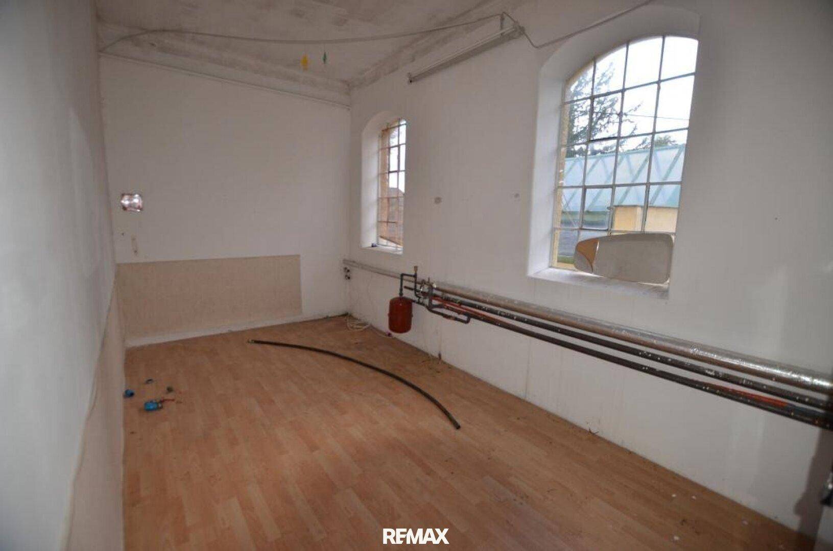 Ein leerer Raum in Renovierung mit großen Bogenfenstern, sichtbaren Rohren und hellem Laminatboden.