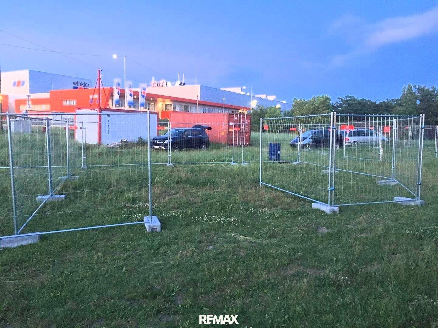 Grasbewachsenes Grundstück mit temporären Metallzäunen und Gewerbegebäuden im Hintergrund unter einem bewölkten Himmel.