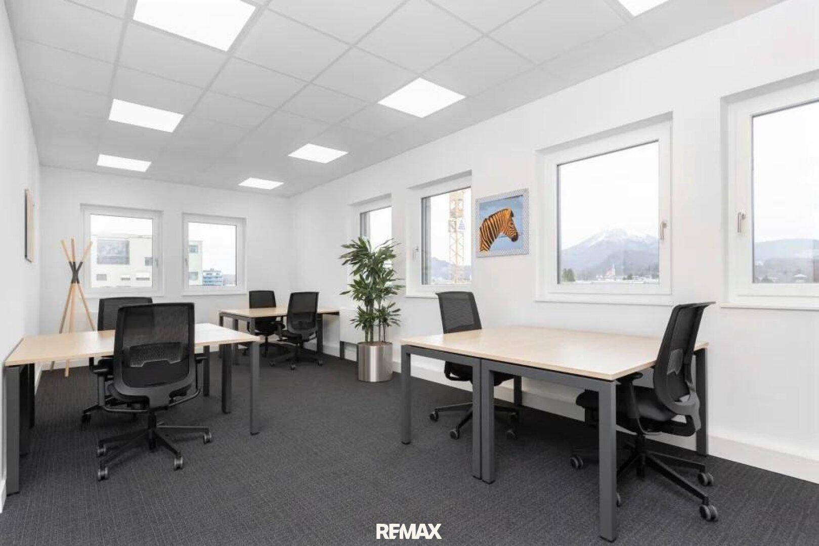 Modernes Büro mit Teppichboden, mehreren Arbeitsplätzen und viel Tageslicht durch große Fenster mit Bergblick.