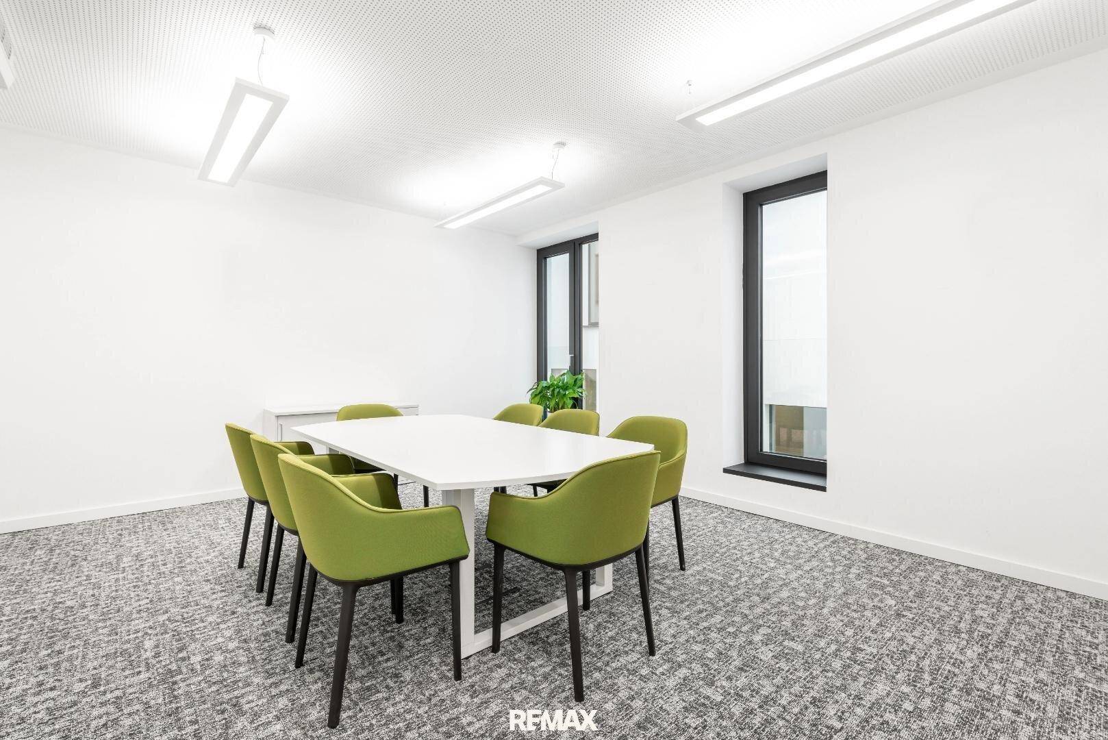 Kleiner Besprechungsraum mit weißem Tisch, grünen Stühlen und Teppichboden.