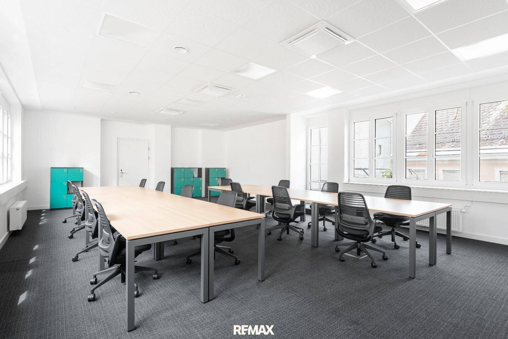 Großes, helles Büro mit mehreren Arbeitsplätzen, Teppichboden und vielen Fenstern für Tageslicht.