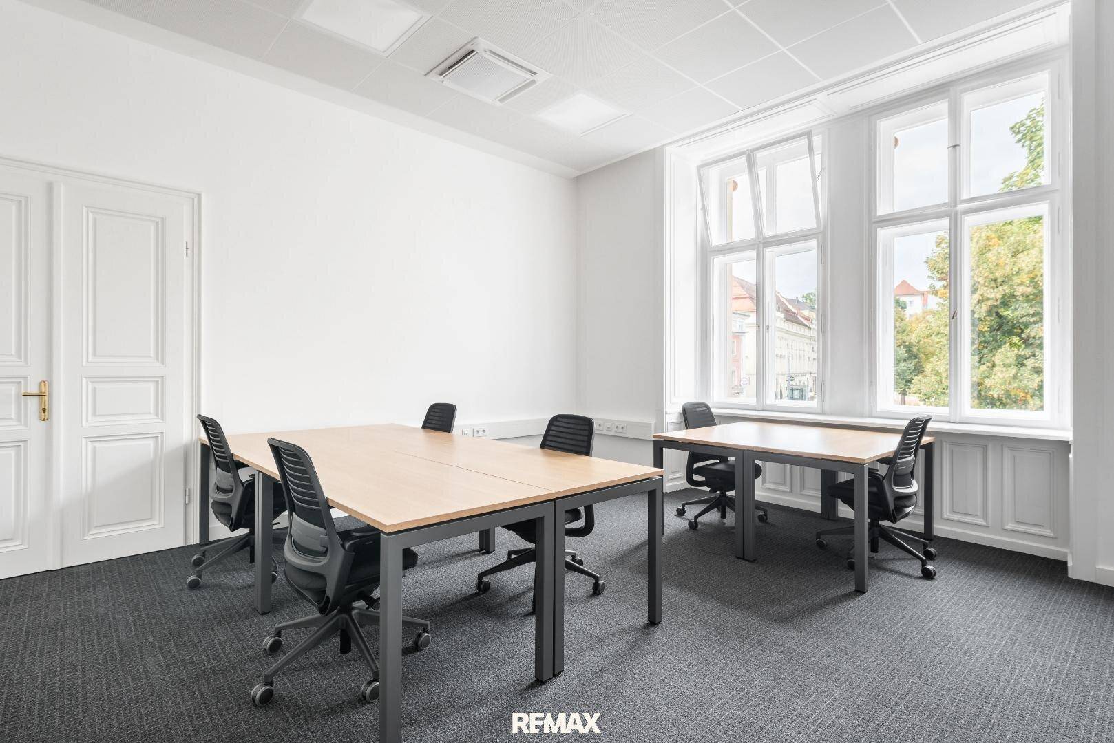 Helles Büro mit mehreren Arbeitsplätzen, grauem Teppichboden und großen Fenstern für eine angenehme Arbeitsatmosphäre.