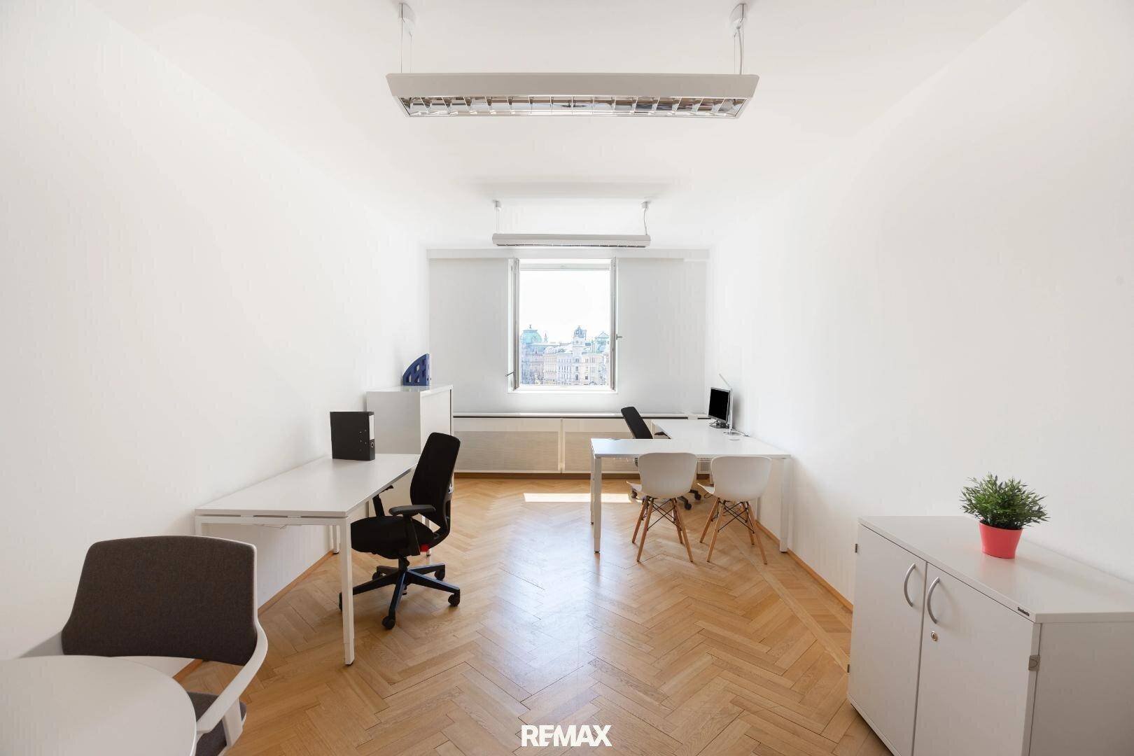 Geräumiges Büro mit mehreren Arbeitsplätzen, einem großen Fenster und hellem Fischgrätparkett für eine angenehme Atmosphäre.