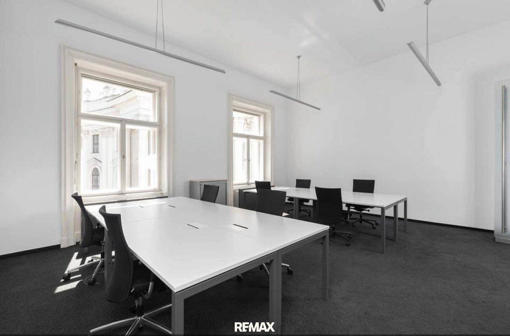 Modernes Büro mit mehreren Arbeitsplätzen, großen Fenstern und dunklem Teppichboden in Schwarz-Weiß-Optik.