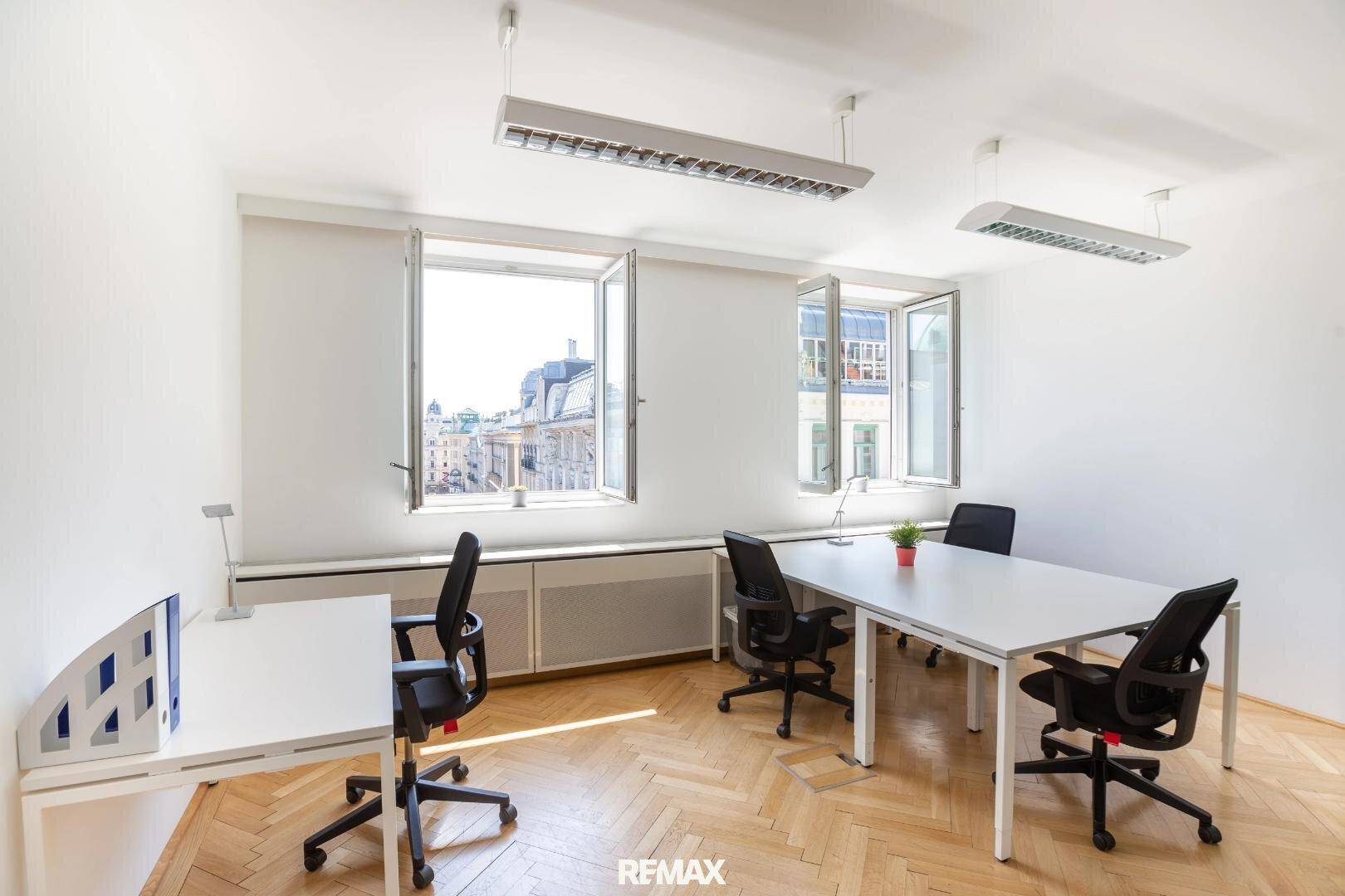 Helles Büro mit mehreren Arbeitsplätzen, großen Fenstern und Fischgrätparkett, das viel Tageslicht hereinlässt.