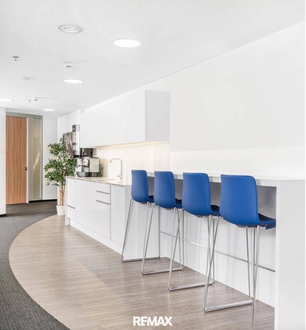 Moderne Büroküche mit weißer Theke, blauen Barhockern, integrierten Geräten und einer Kombination aus Laminat- und Teppichboden.