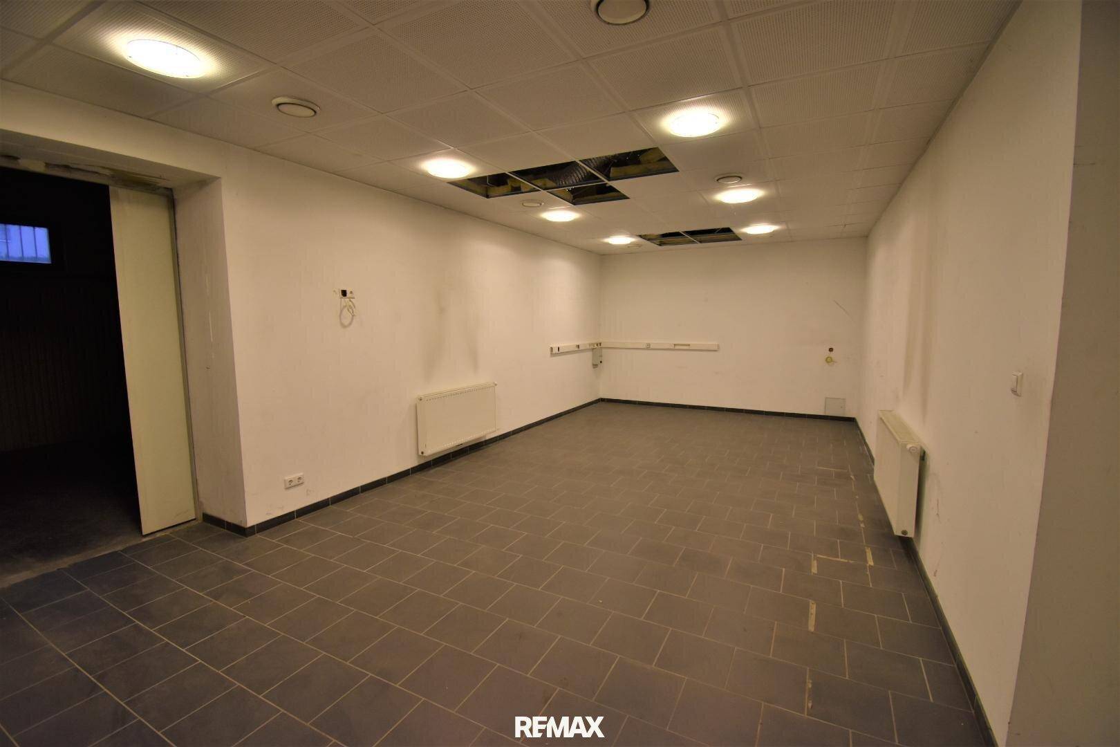 Heller, leerer Raum mit dunklen Fliesen, weißen Wänden und abgehängter Decke mit Einbauleuchten.