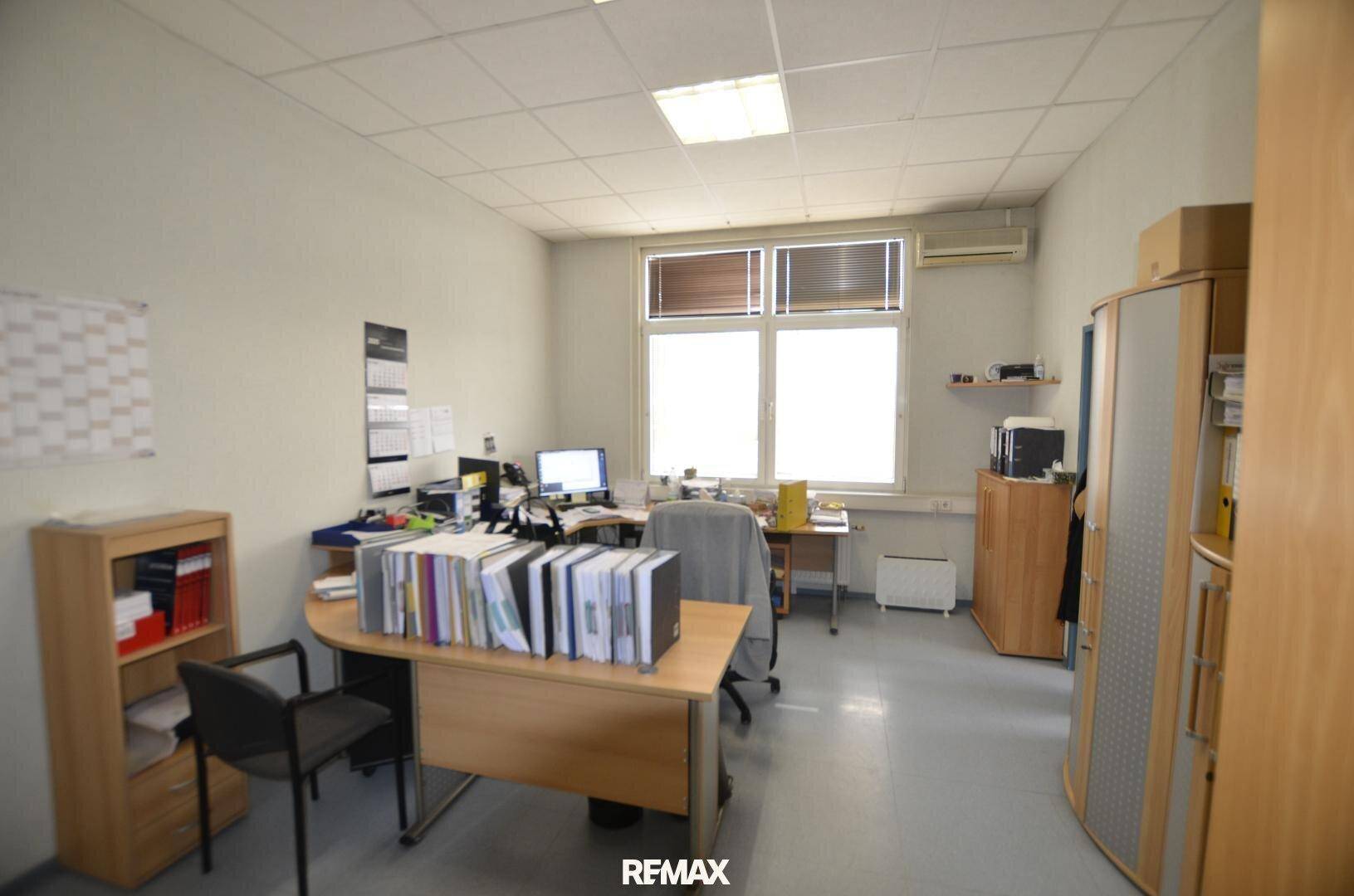 Funktionales Büro mit Schreibtischen, Bürostühlen und Holzschränken, beleuchtet durch ein großes Fenster.