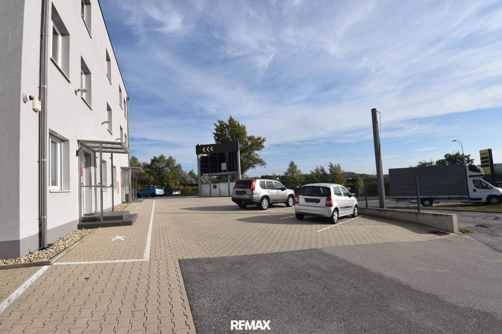 Außenansicht eines modernen Gebäudes mit Parkplätzen und Bäumen im Hintergrund.