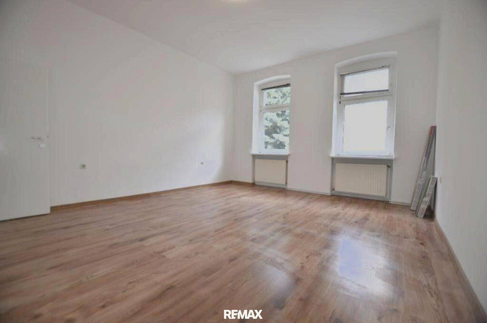Leeres Zimmer mit Laminatboden und zwei Fenstern, bereit zur individuellen Gestaltung.