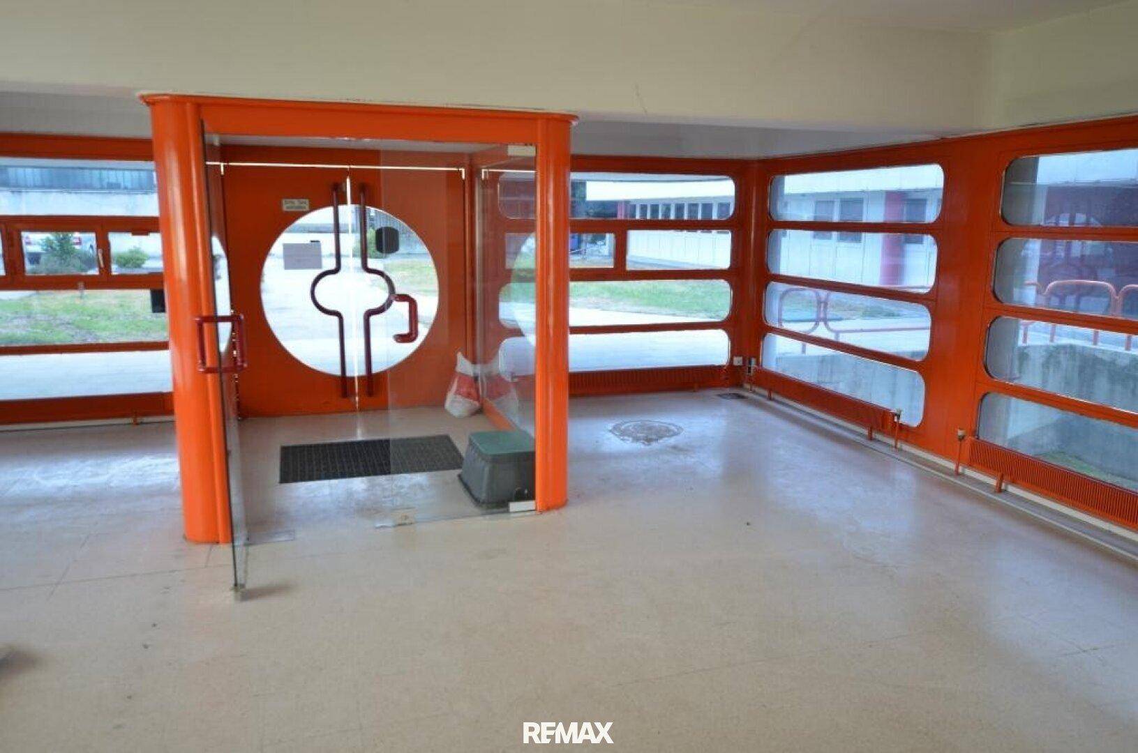 Eingangsbereich mit markanter runder Glastür, Fliesenboden und orangefarbenen Rahmen.