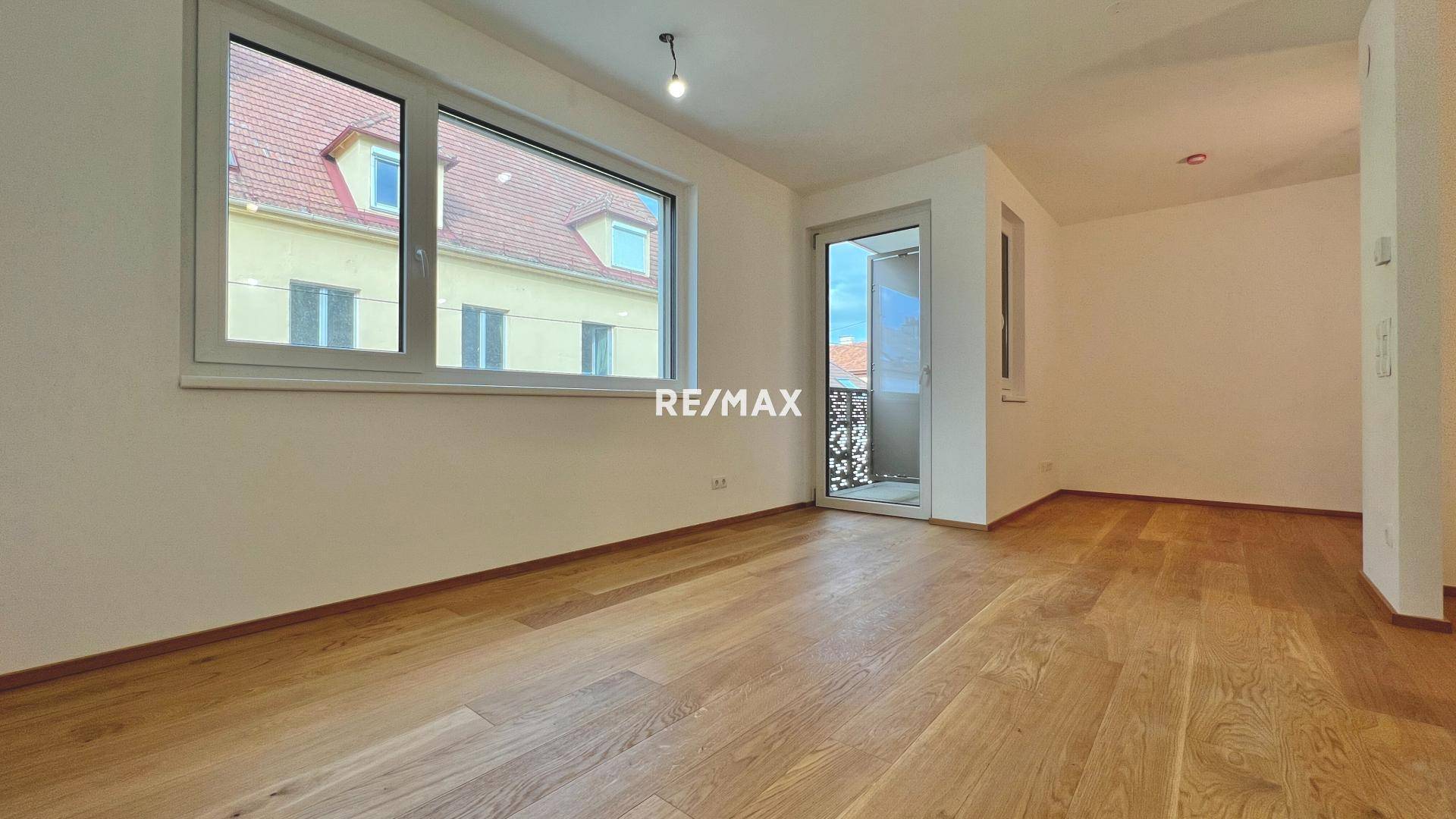 Lichtdurchfluteter Wohnbereich mit großen Fenstern, Holzboden und Zugang zu einem kleinen Balkon mit Ausblick.