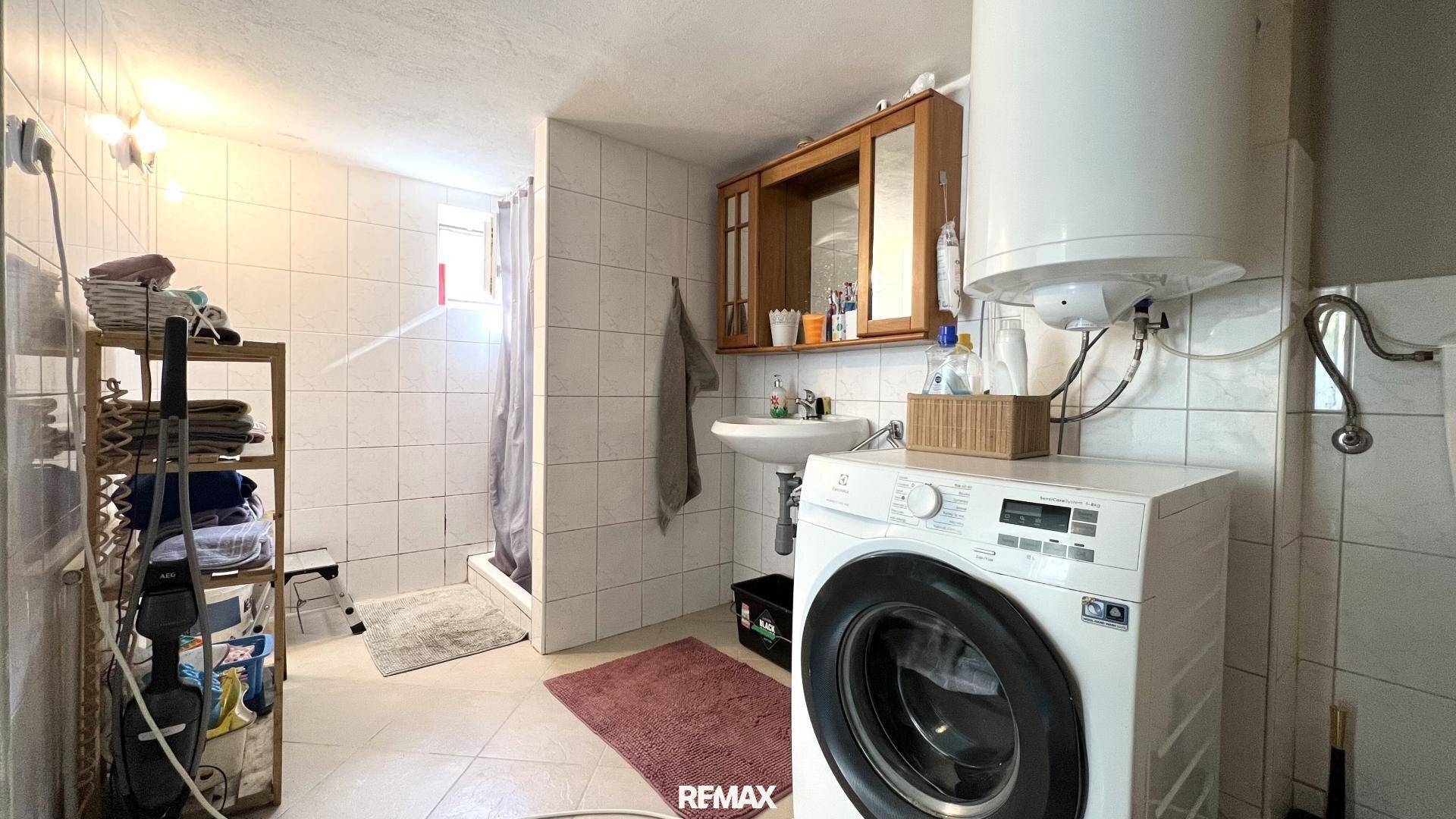 Gefliestes Badezimmer mit Duschkabine, Waschbecken, Waschmaschine und Warmwasserboiler.