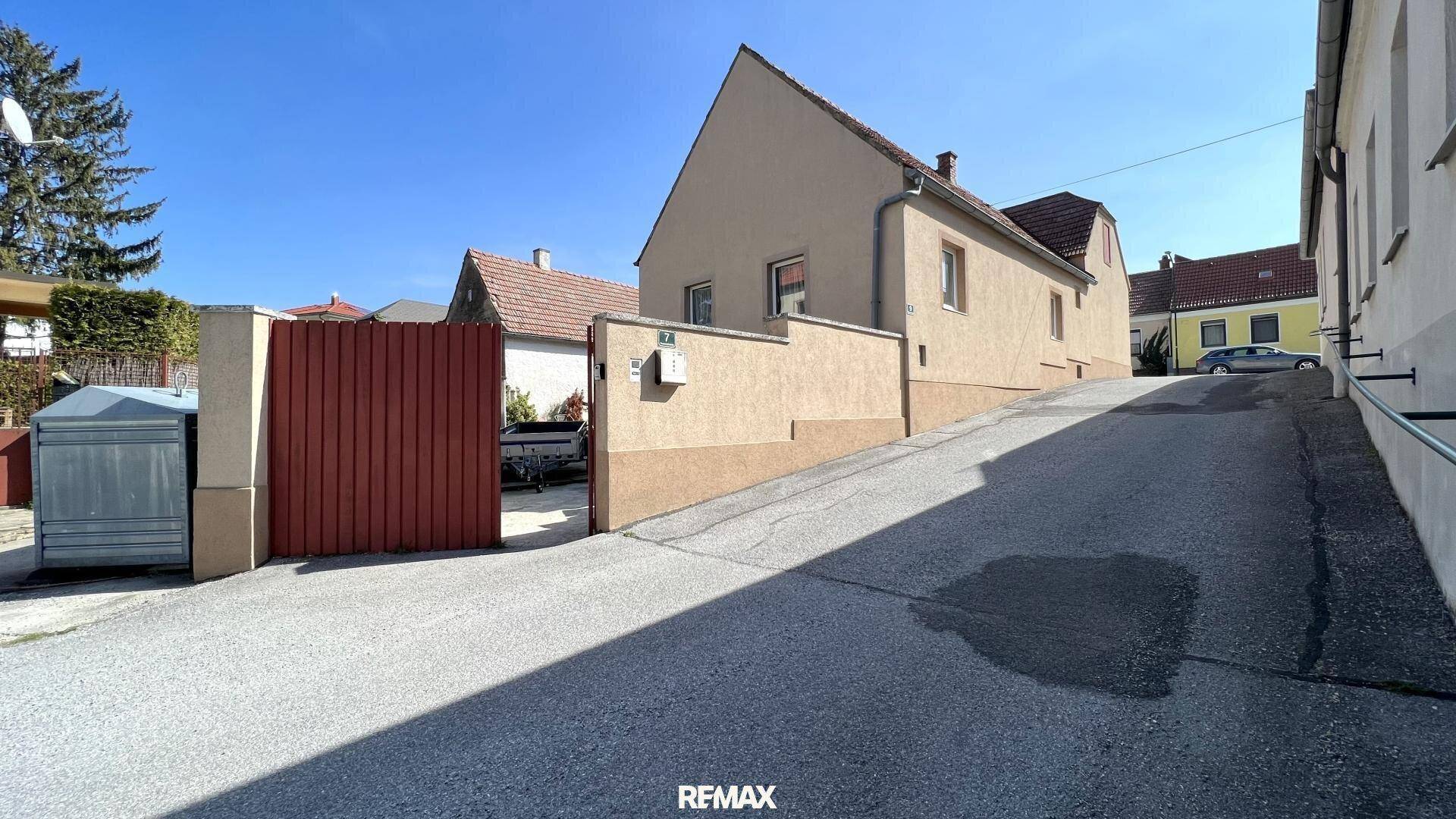 Ansicht eines Hauses von einer leicht ansteigenden Straße mit rotem Tor und kleiner Mauer.