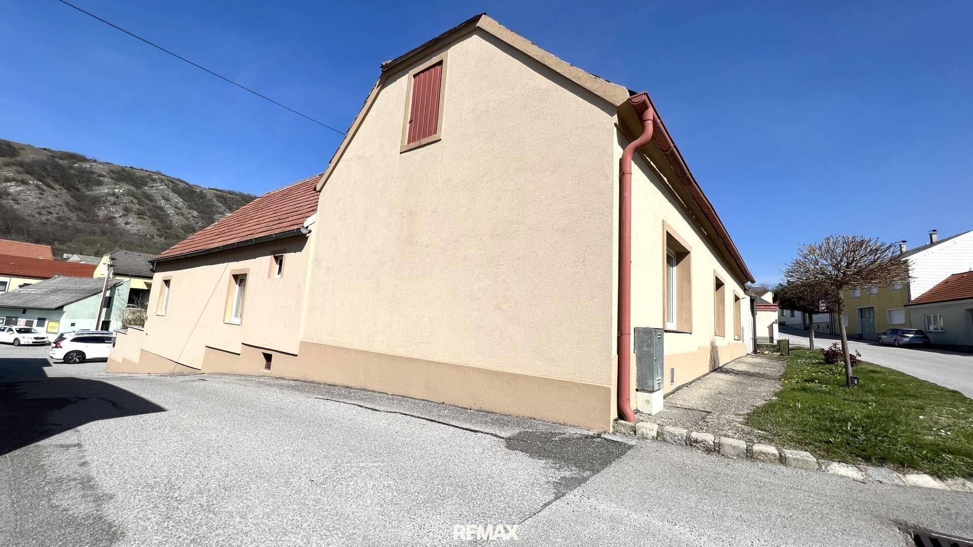 Seitenansicht eines Einfamilienhauses mit beiger Fassade und rotem Dach in ländlicher Umgebung.