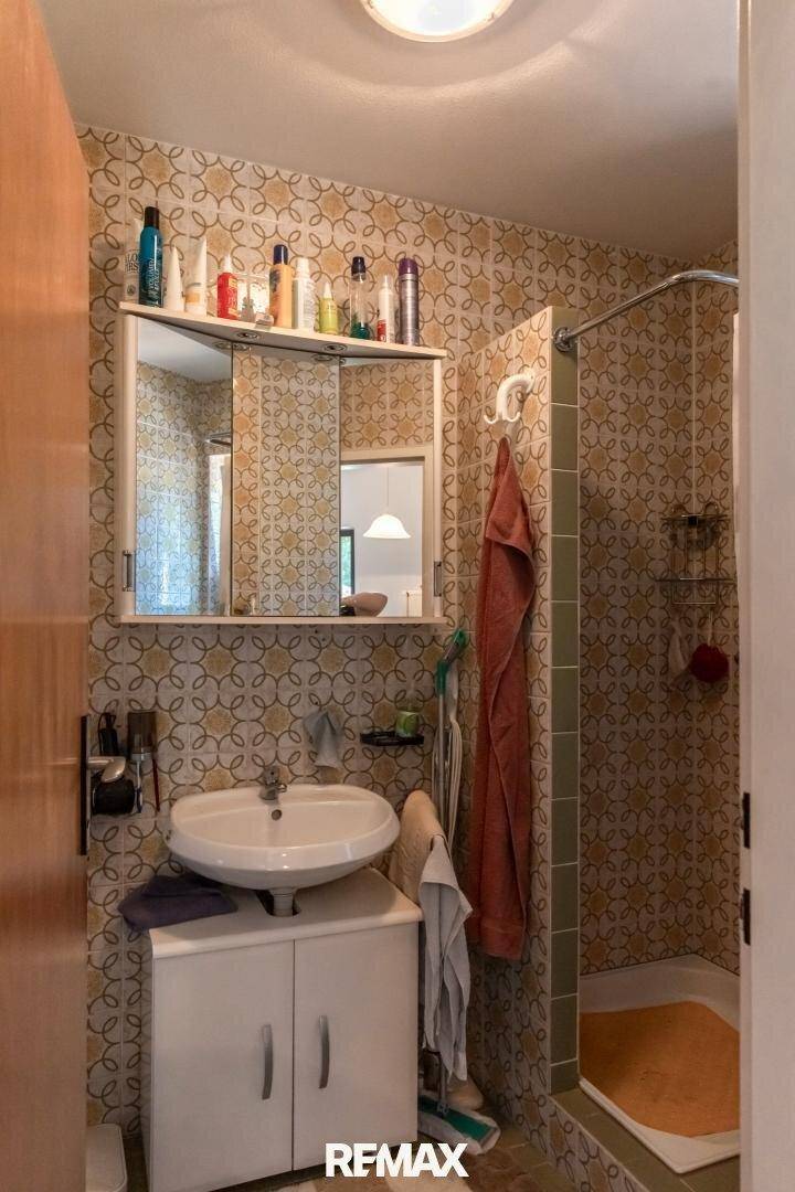 Badezimmer mit Waschbecken, Spiegelschrank und Duschkabine, ausgestattet mit gemusterten Fliesen.