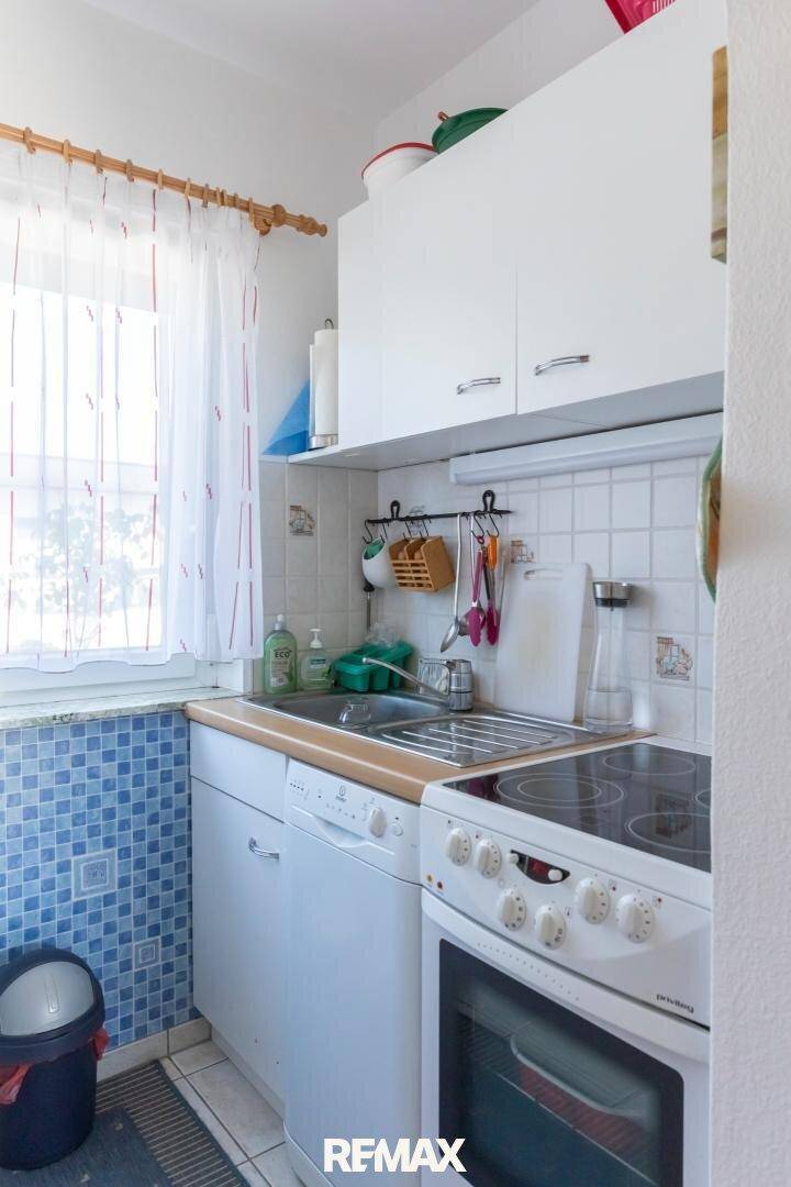 Kleine Küche mit weißen Schränken, Spüle und Elektroherd, beleuchtet durch ein Fenster.