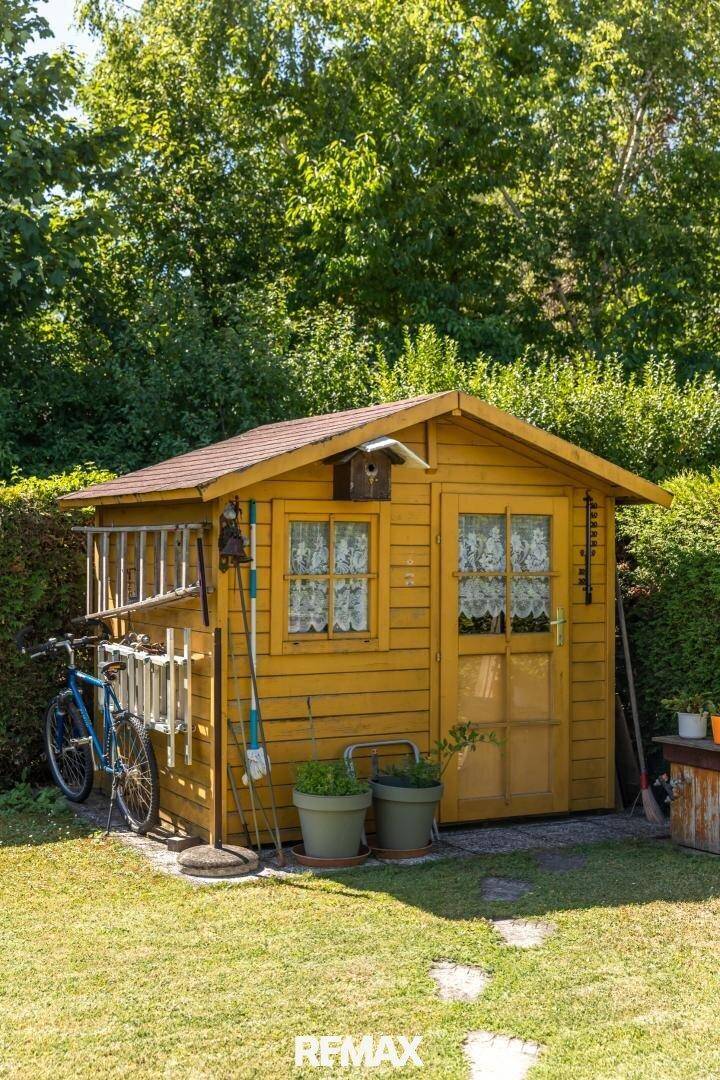 Gelber Holzschuppen im Garten, umgeben von Grünflächen und Bäumen, mit Gartengeräten.