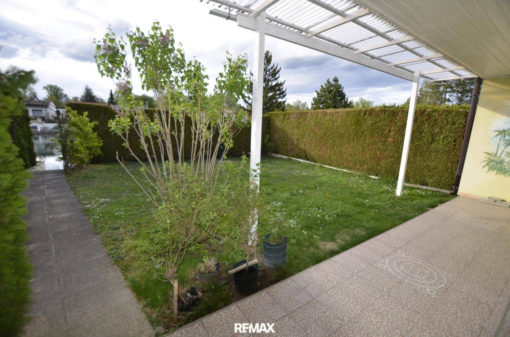 Gartenbereich mit Rasenfläche, gepflastertem Weg und überdachter Terrasse, umgeben von Hecken.