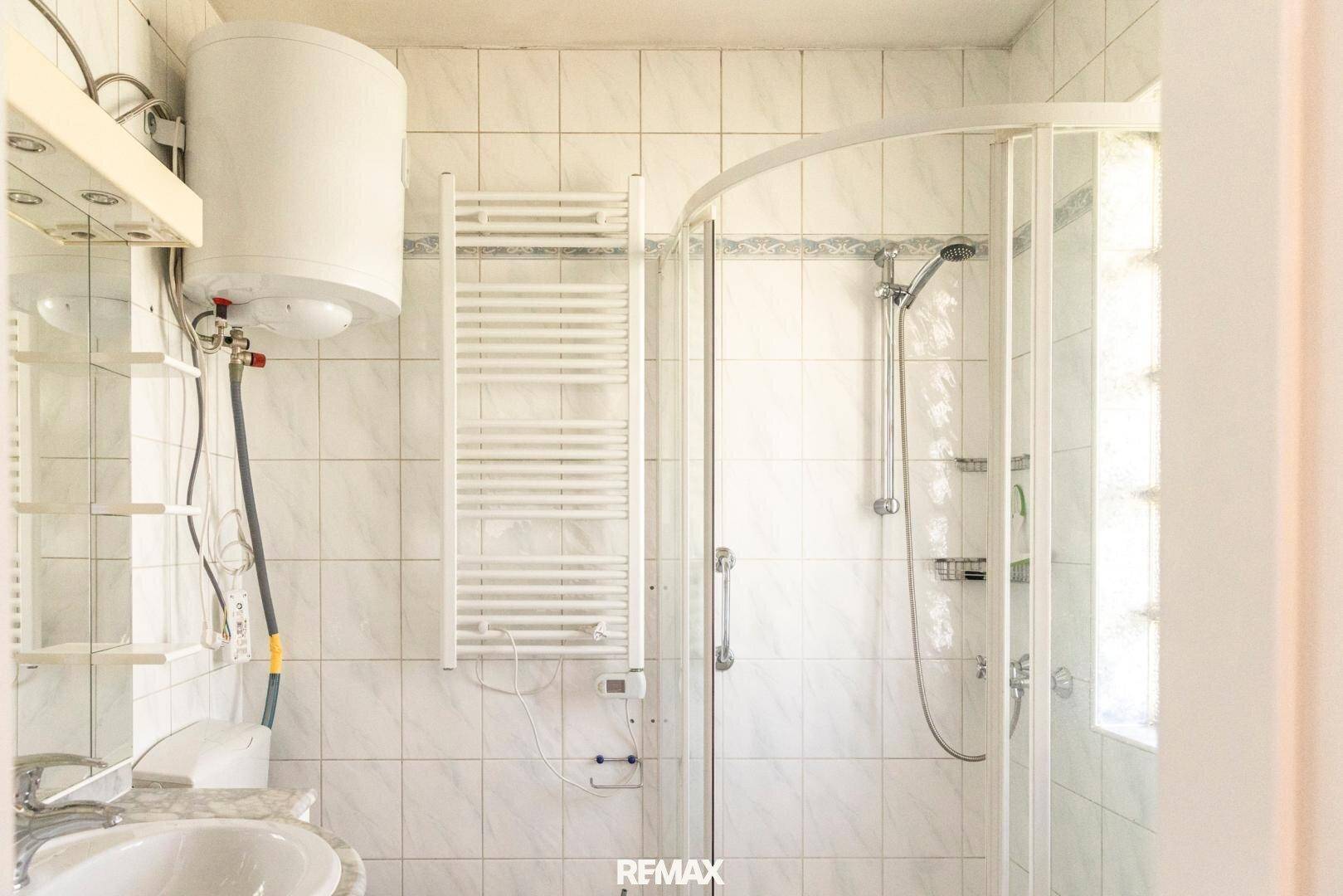 Badezimmer mit Duschkabine, Waschbecken, Handtuchheizkörper und Warmwasserboiler, gefliest.