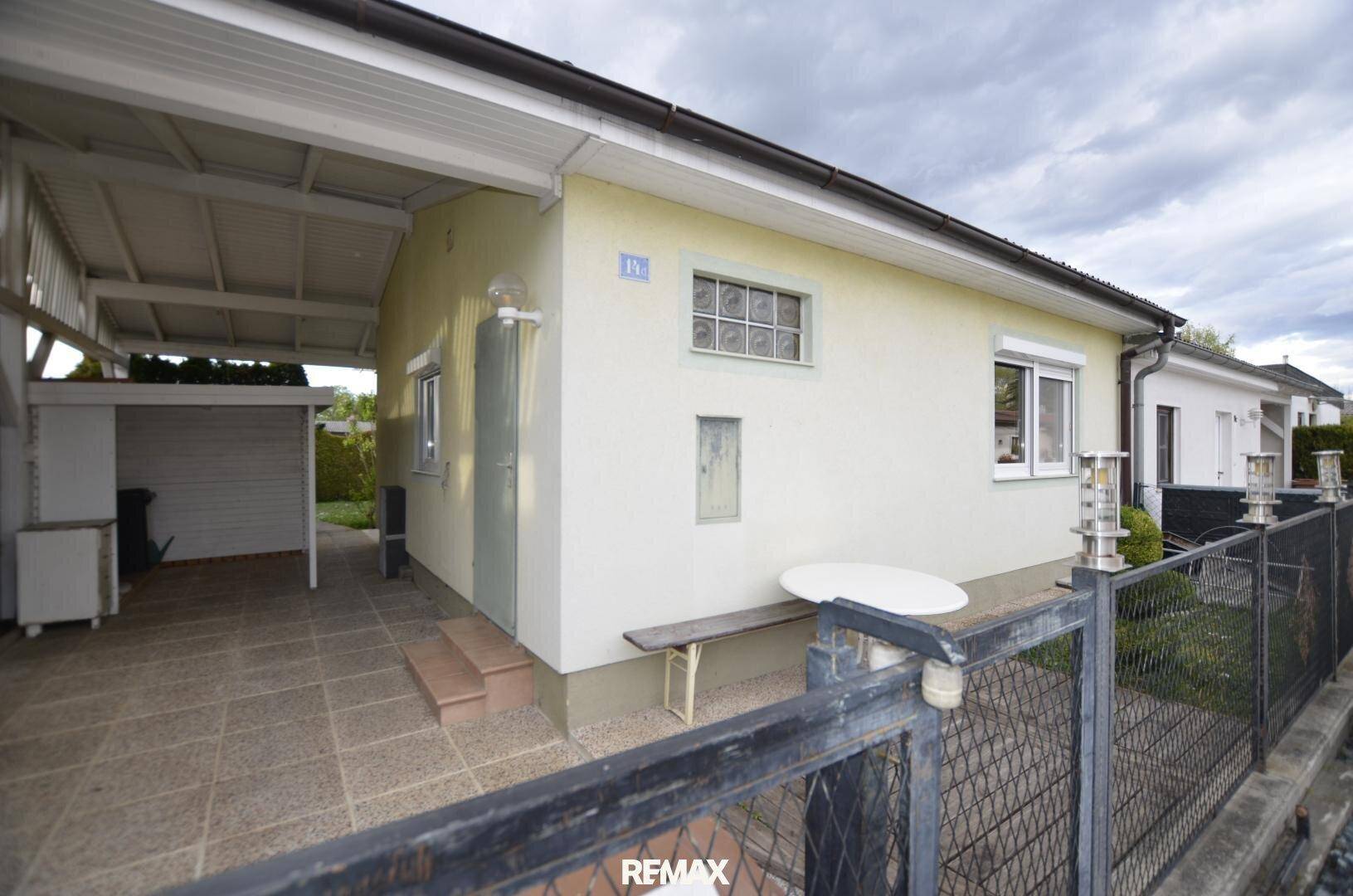 Seitenansicht des Hauses mit überdachtem Bereich und gepflastertem Vorplatz, umzäunt.