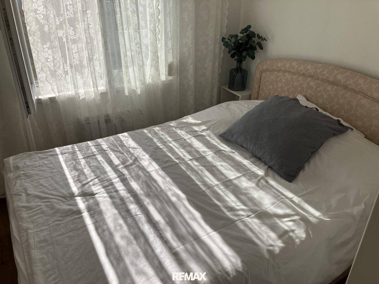Helles Schlafzimmer mit Einzelbett und Spitzenvorhängen, durch die Sonnenlicht fällt.