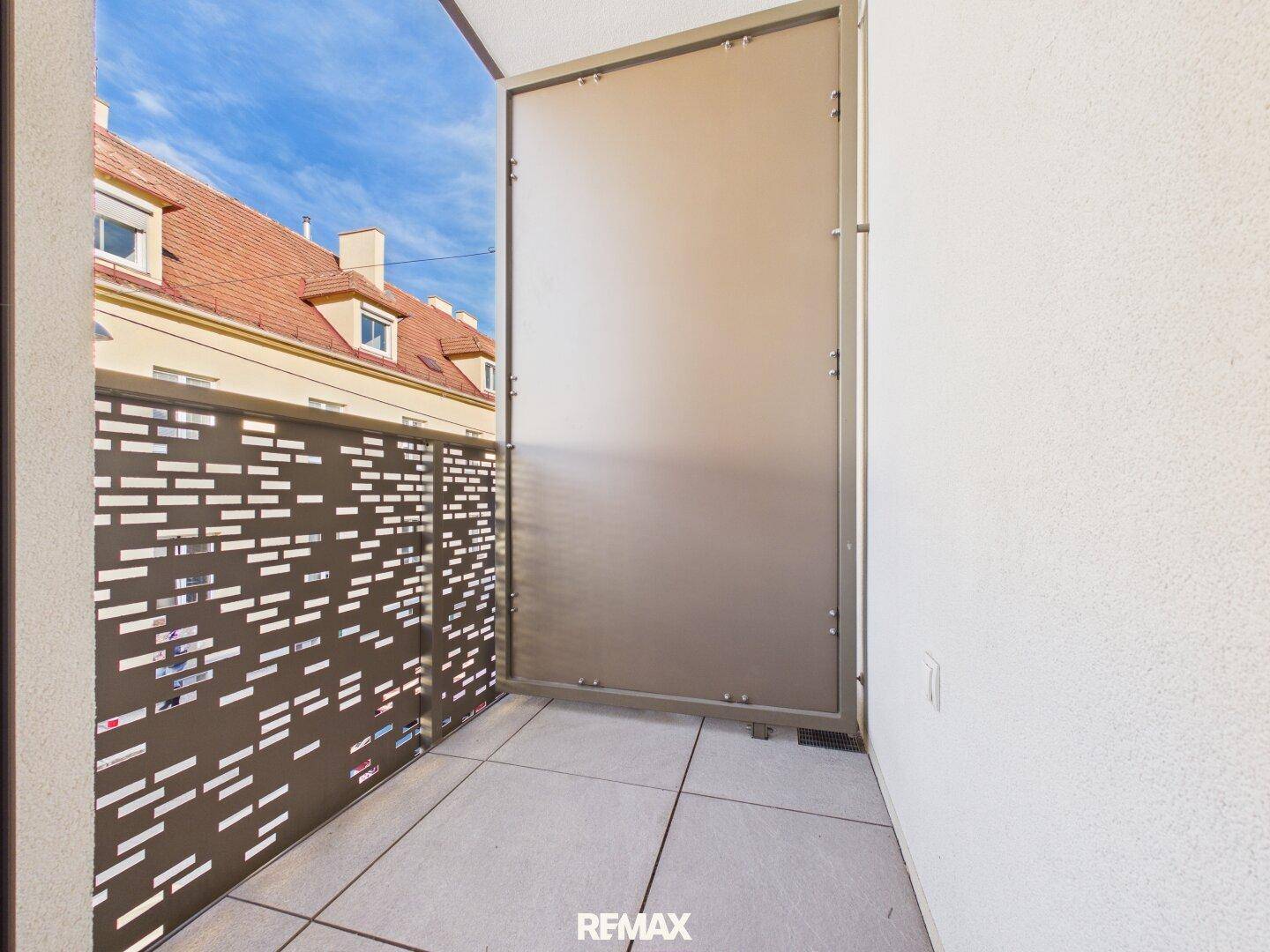 Moderner Balkon mit Fliesenboden und perforierter Metallbrüstung unter blauem Himmel.