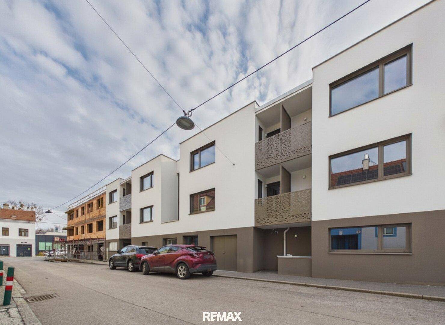 Moderne Wohngebäude mit Balkonen und Parkplätzen an einer ruhigen Straße unter bewölktem Himmel.