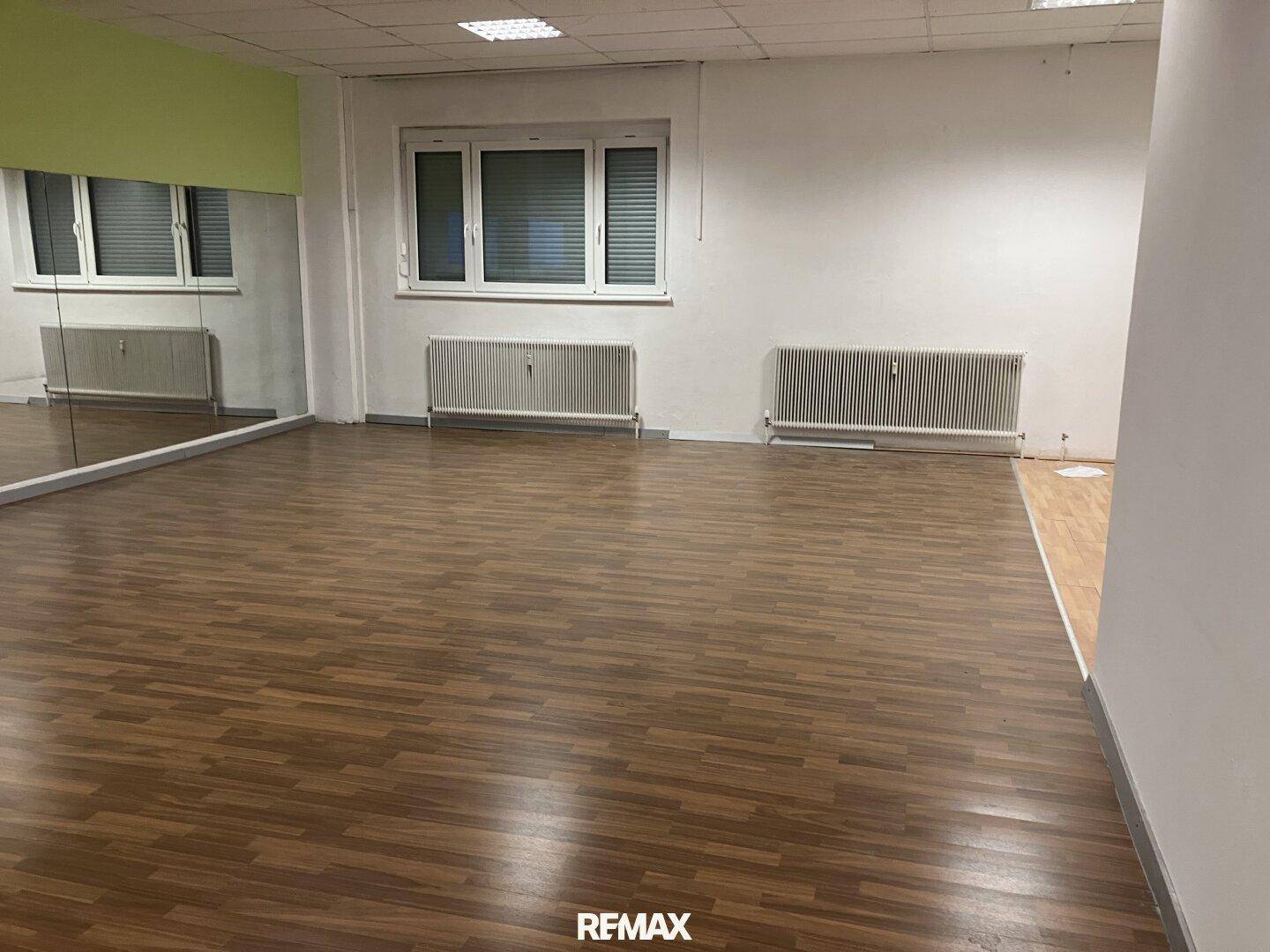 Leerer Mehrzweckraum mit Holzfußboden, großen Fenstern, Heizkörpern und einer Spiegelwand.