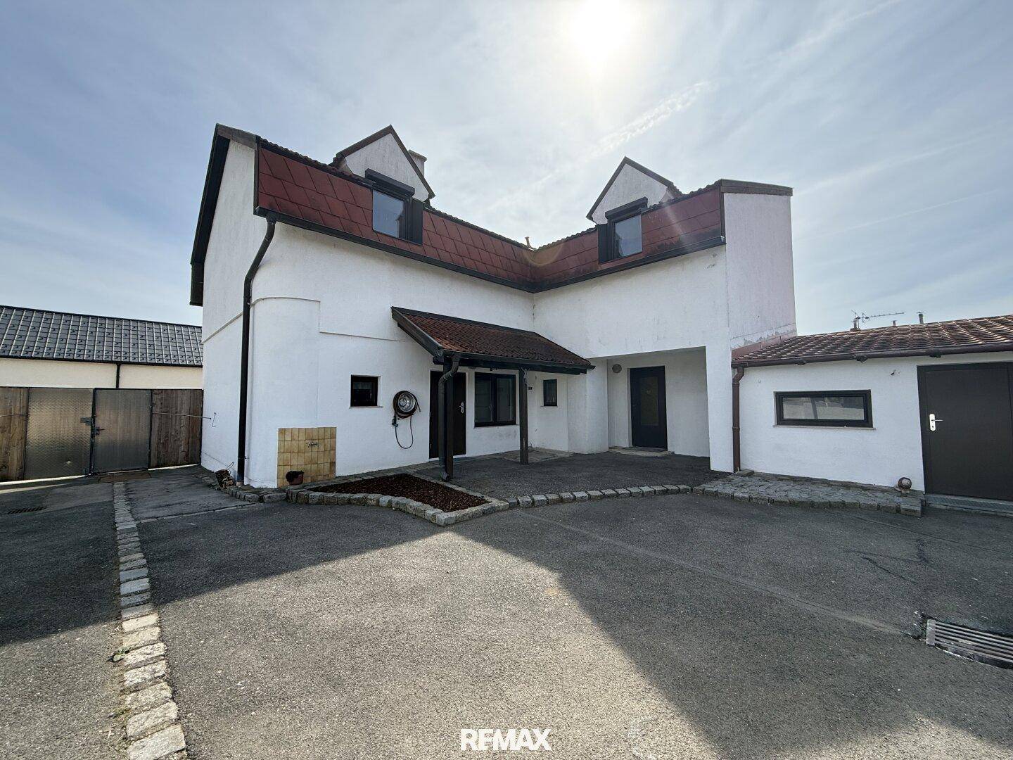 Weißes Einfamilienhaus mit rotem Dach und gepflasterter Auffahrt unter blauem Himmel.