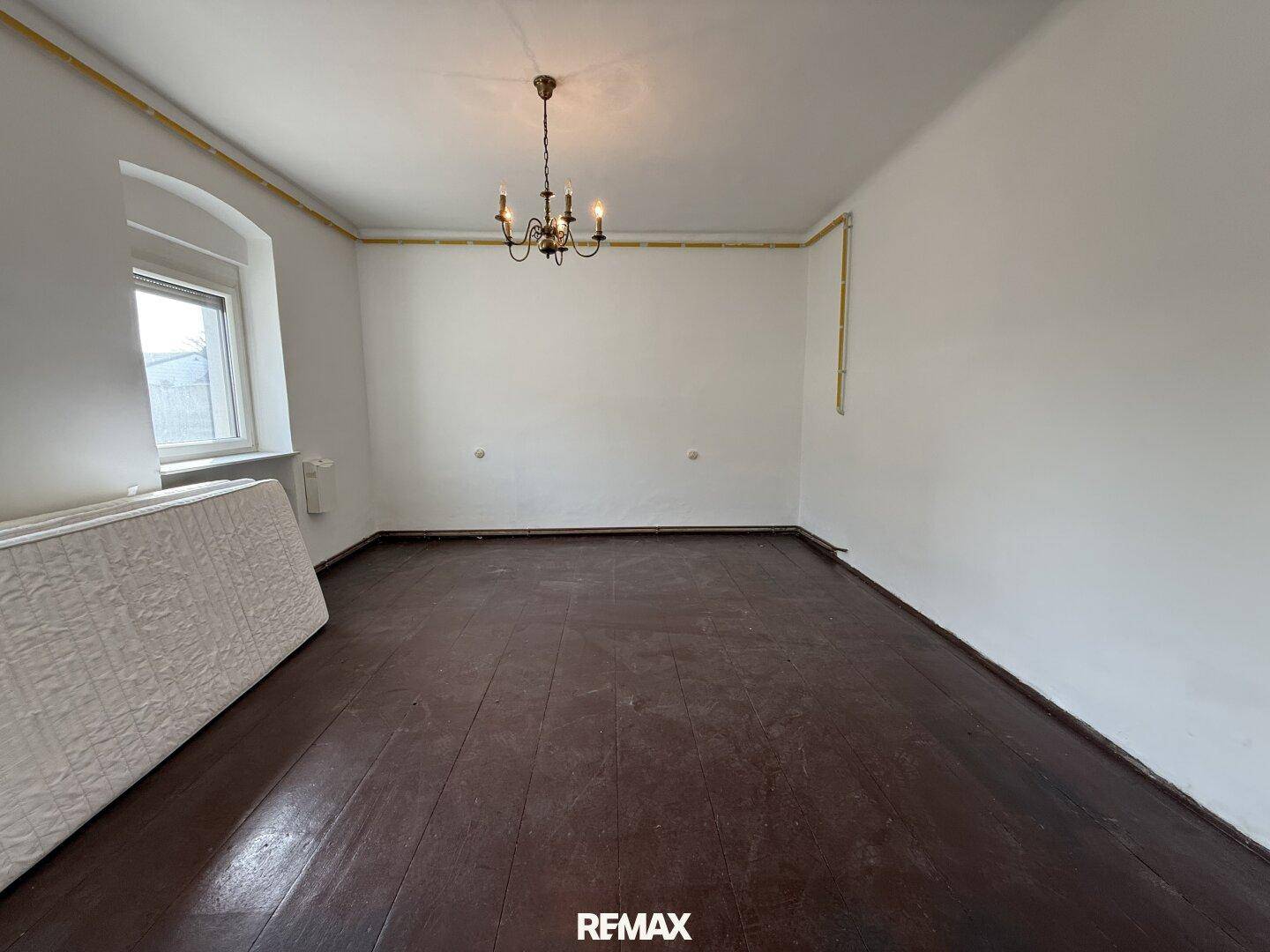 Geräumiges, leeres Zimmer mit dunklem Holzboden, Bogenfenster und Kronleuchter.