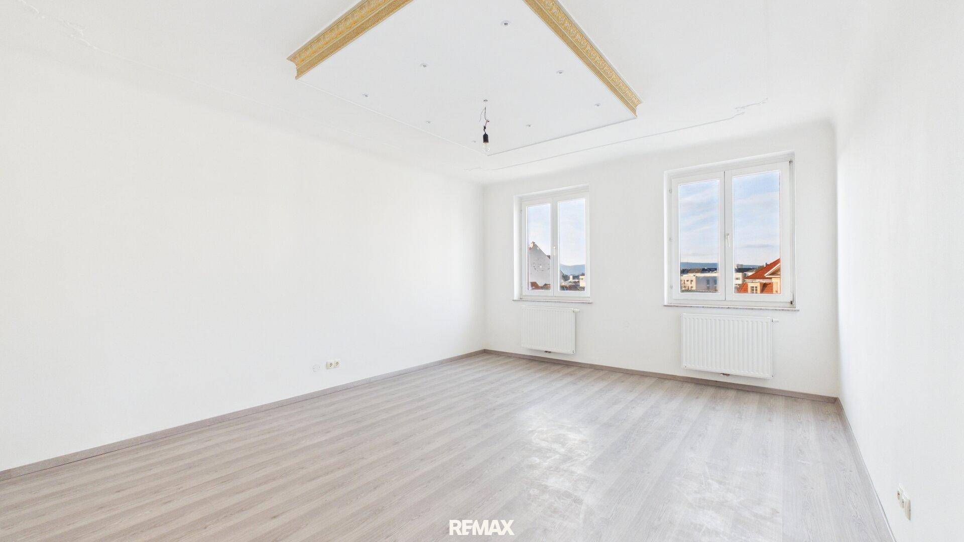 Geräumiges, helles Zimmer mit zwei Fenstern, hellem Laminatboden und einer dekorativen Deckenleiste.