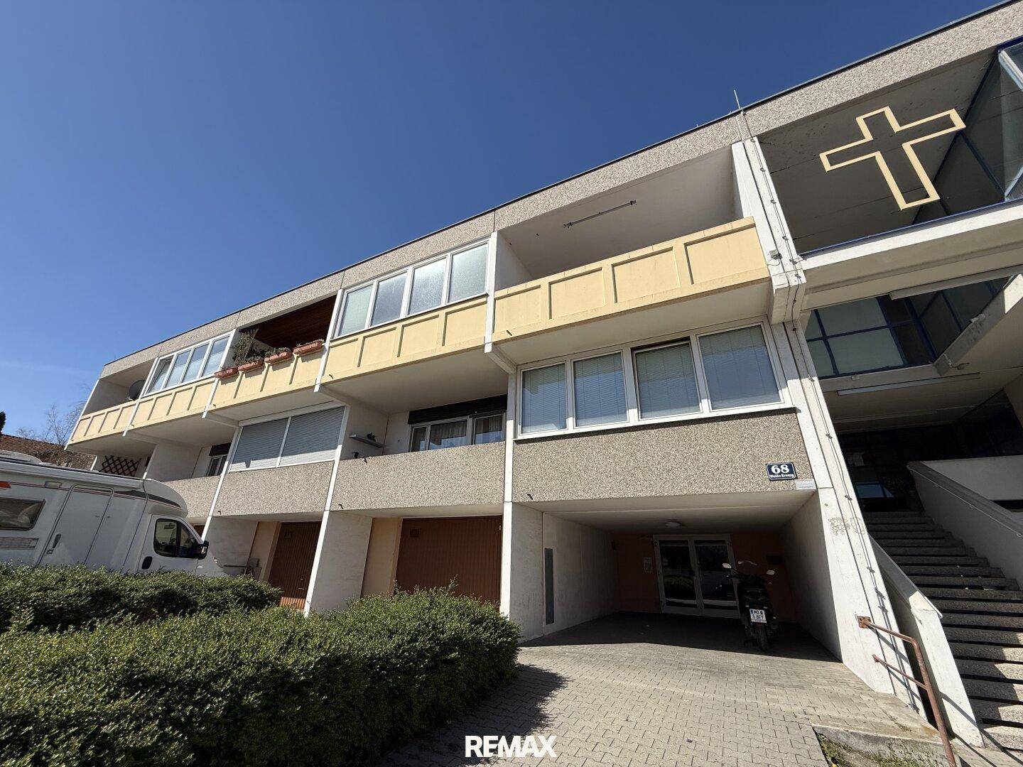 Eingangsbereich eines Mehrfamilienhauses mit Garagenzufahrt und Treppenaufgang unter blauem Himmel.