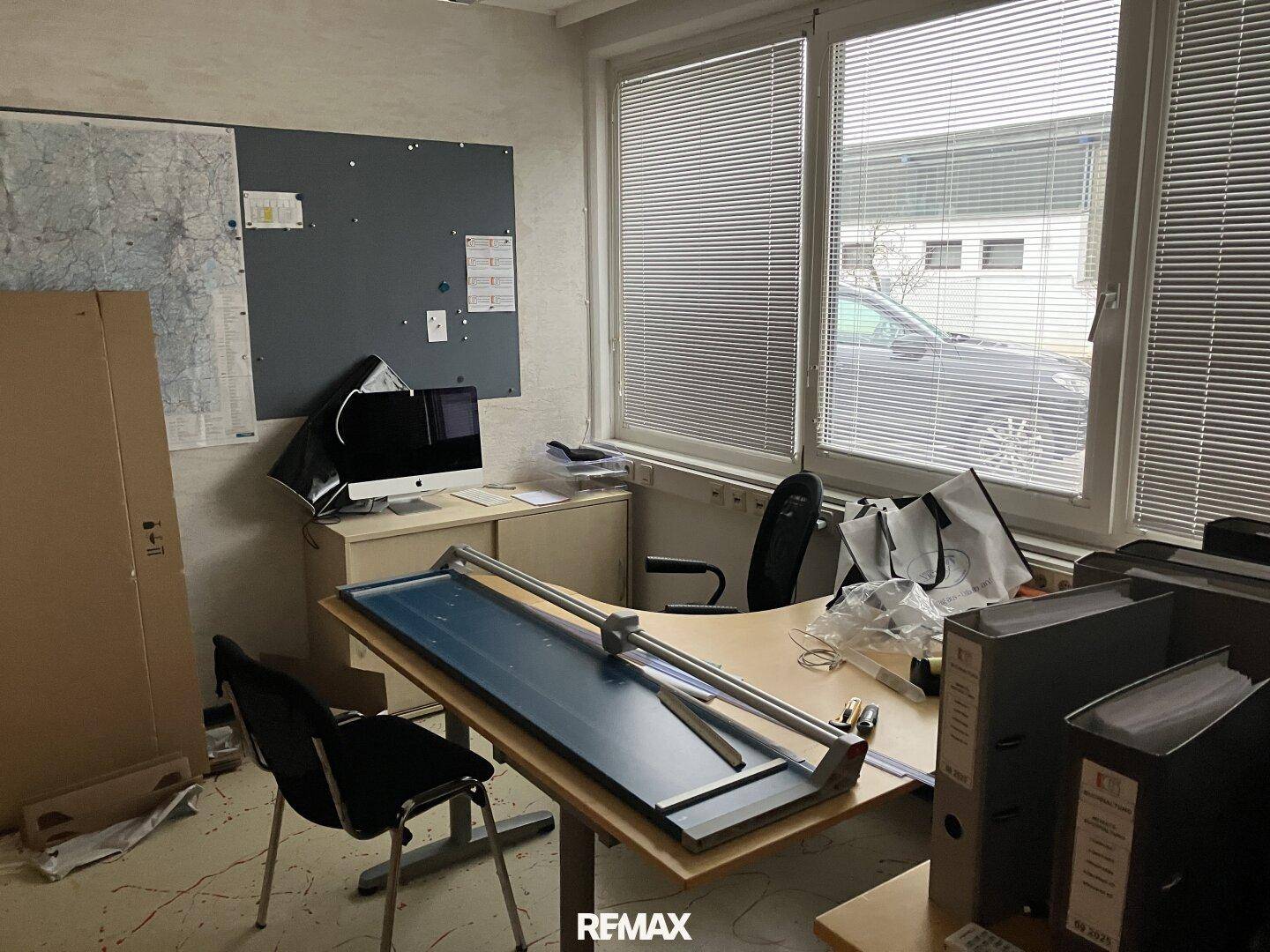 Ein Büroraum mit einem Schreibtisch, Computer, Jalousien am großen Fenster und einer Pinnwand.