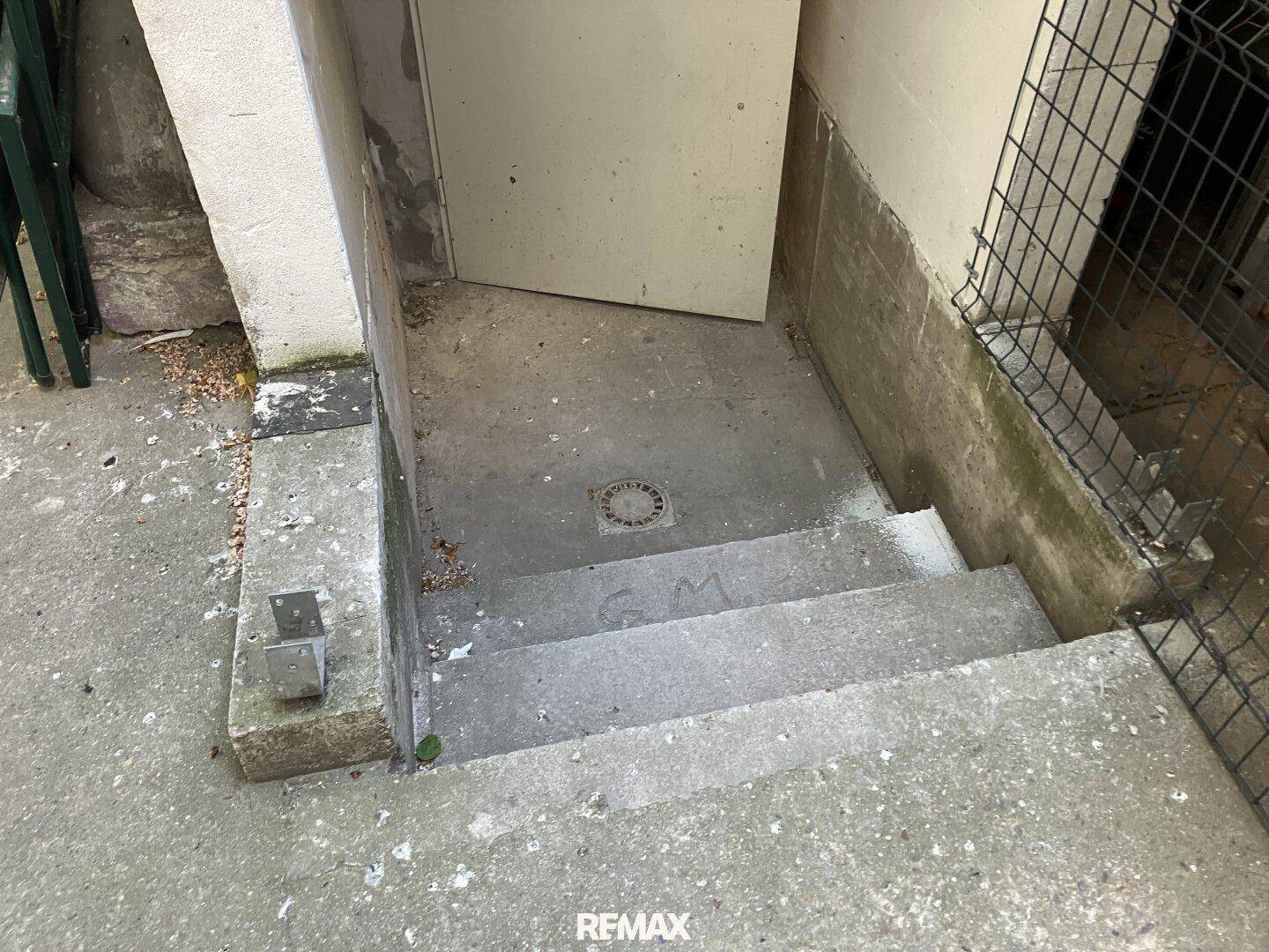 Betonstufen führen zu einem Kellereingang mit einem Abfluss auf dem Podest, umgeben von rauen Wänden.