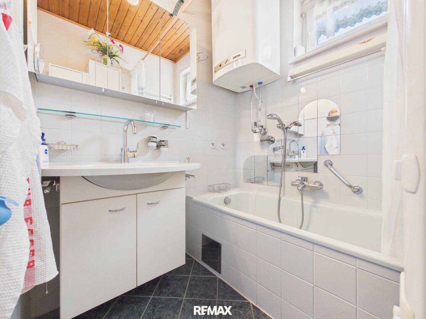 Helles Badezimmer mit Badewanne, Waschbecken, Haltegriffen und einem Fenster.