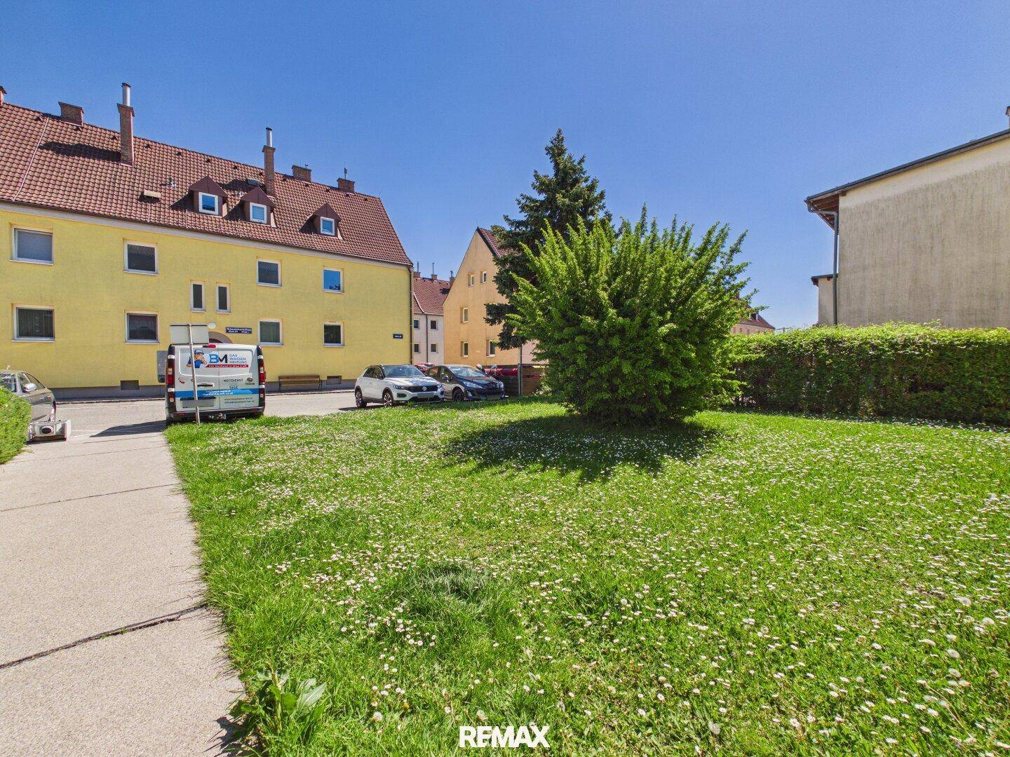 Grüne Rasenfläche mit Parkplätzen und Wohngebäuden im Hintergrund unter blauem Himmel.