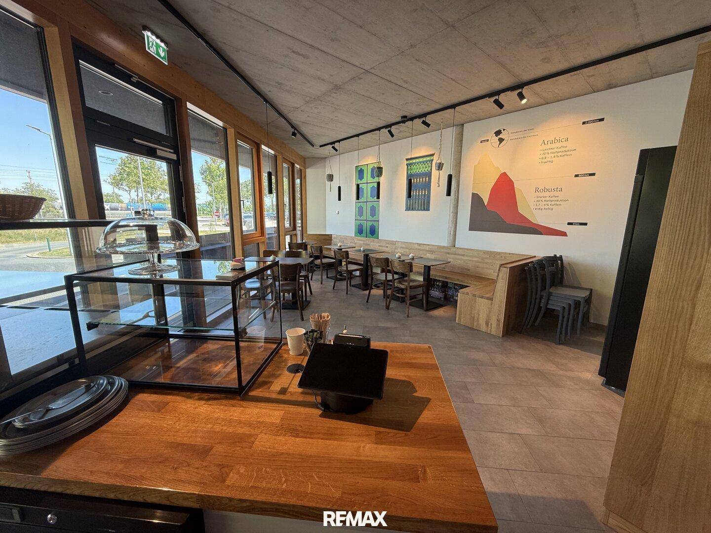 Gemütlicher Sitzbereich eines Cafés mit Holztischen, Stühlen und einer langen Bank, hell durch große Fenster.
