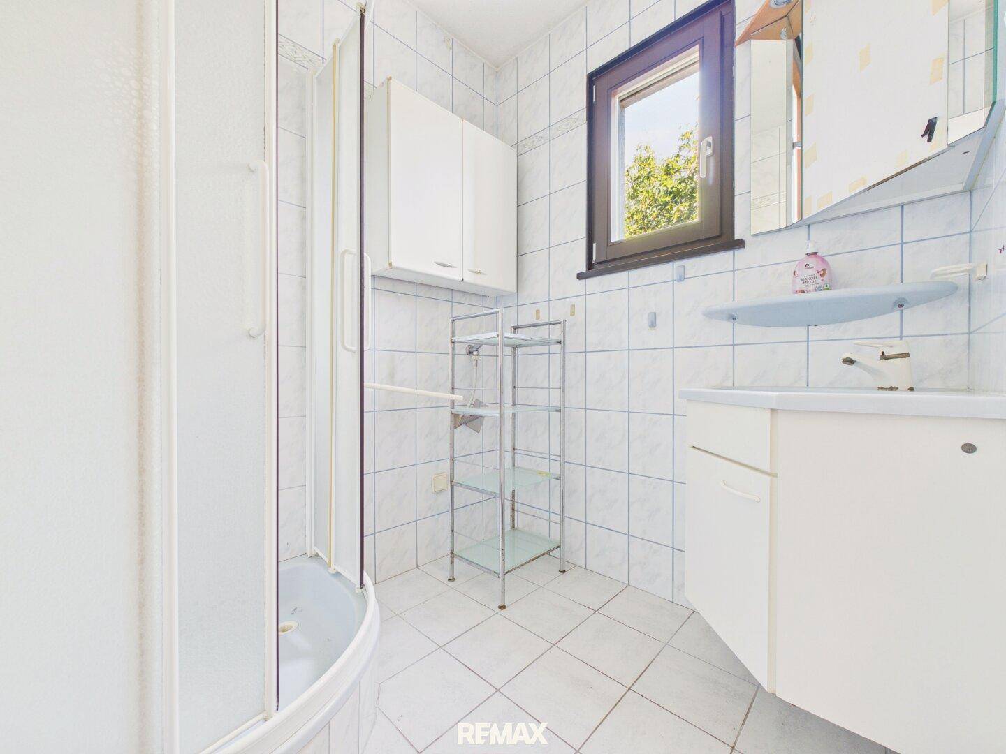 Gefliestes Badezimmer im 80er-Jahre-Stil mit Duschkabine, Waschbecken und Fenster.