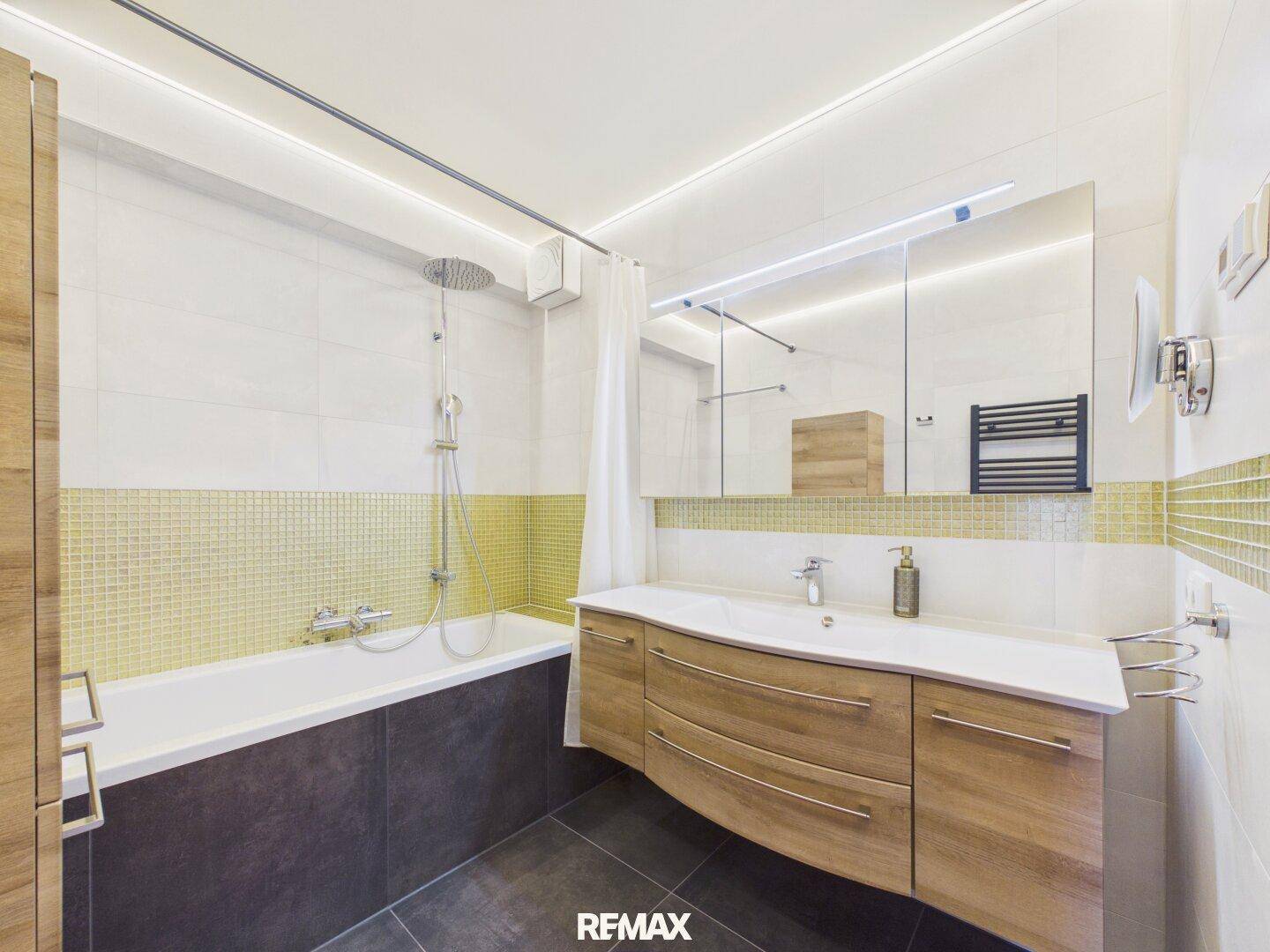 Stilvolles Badezimmer mit integrierter Badewanne und Dusche, Holzwaschtisch und goldfarbenen Akzentfliesen.