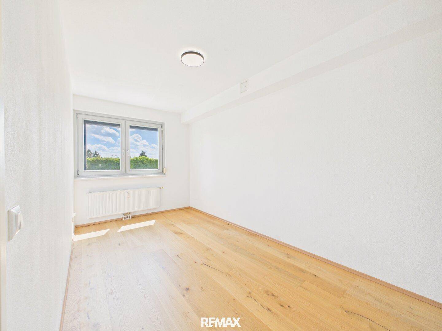 Helles, leeres Zimmer mit Holzboden, großem Fenster und Heizkörper, ideal als Schlaf- oder Arbeitszimmer.
